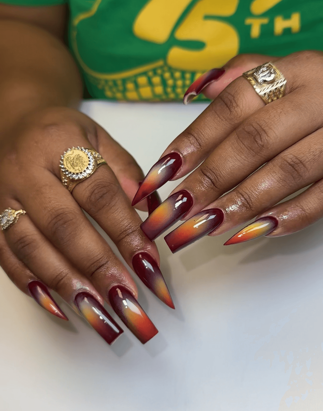Hands with long, decorated nails and multiple rings, resting on a white surface, with a green and yellow shirt in background.