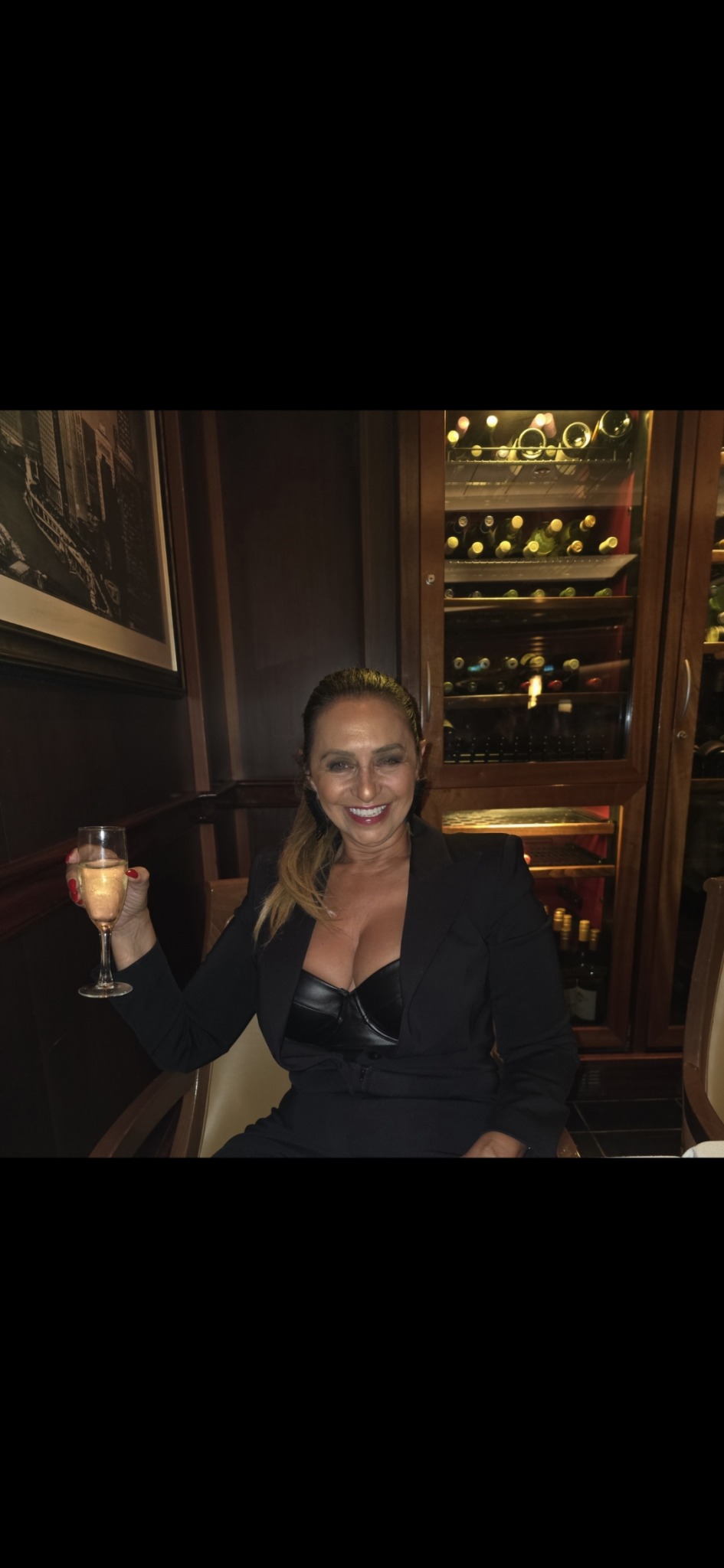 Woman holding a drink in a wine cellar or bar setting, smiling, with wine bottles behind her.