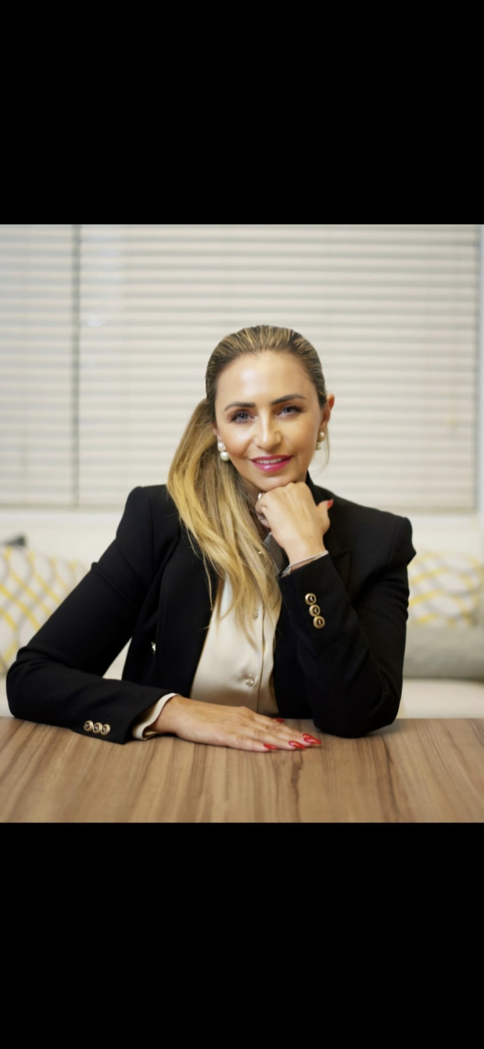 Woman with long hair in a black blazer sitting at a table, smiling, with blinds in the background.