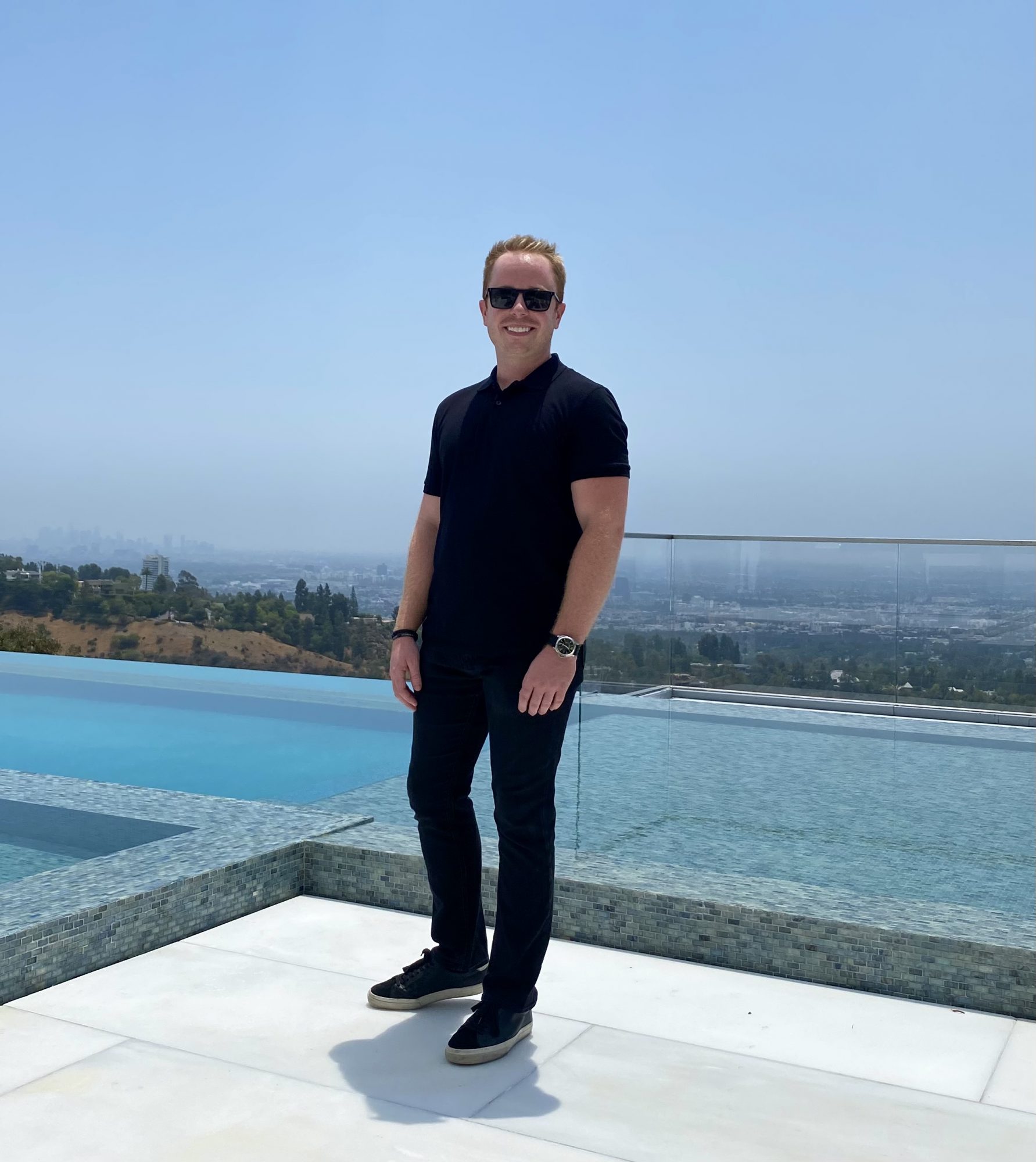 Man standing outdoors on a white surface with a city view and pool behind him, wearing sunglasses, black shirt, and jeans.