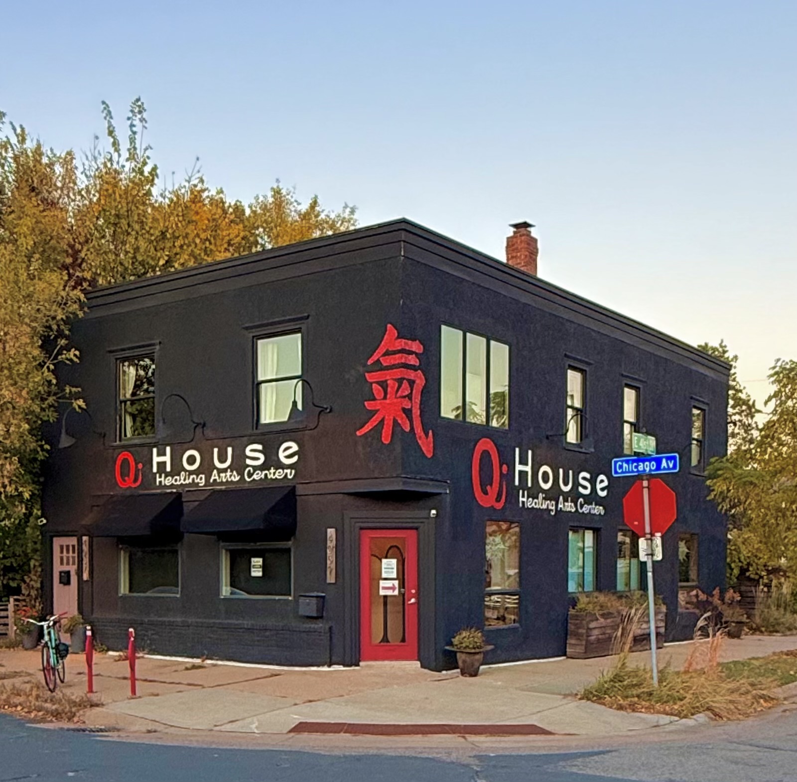 Black two-story building with red and white signage, corner location, trees in background, street signs nearby.