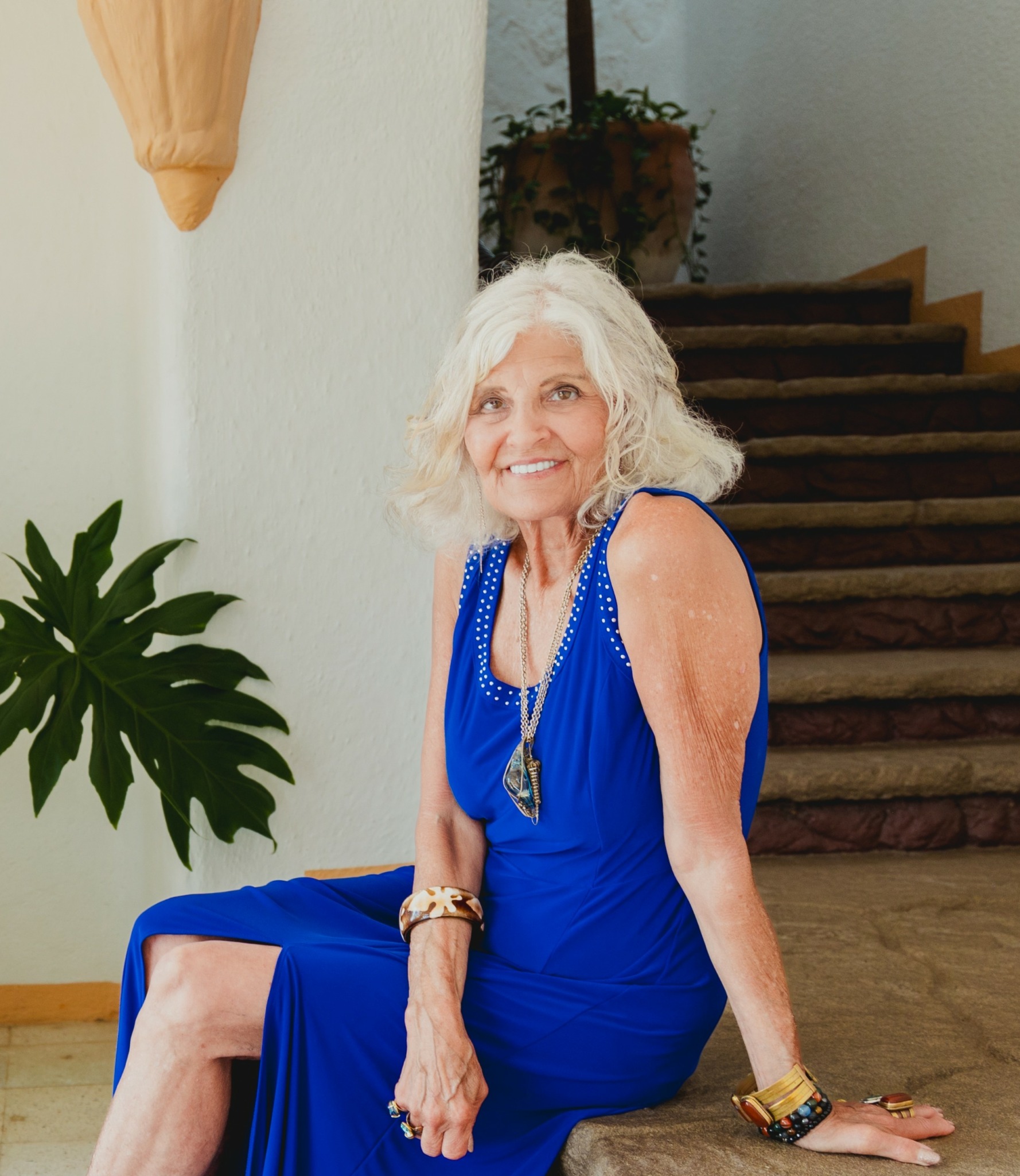 Older woman with curly white hair sitting on stairs, smiling, wearing a blue dress and jewelry.