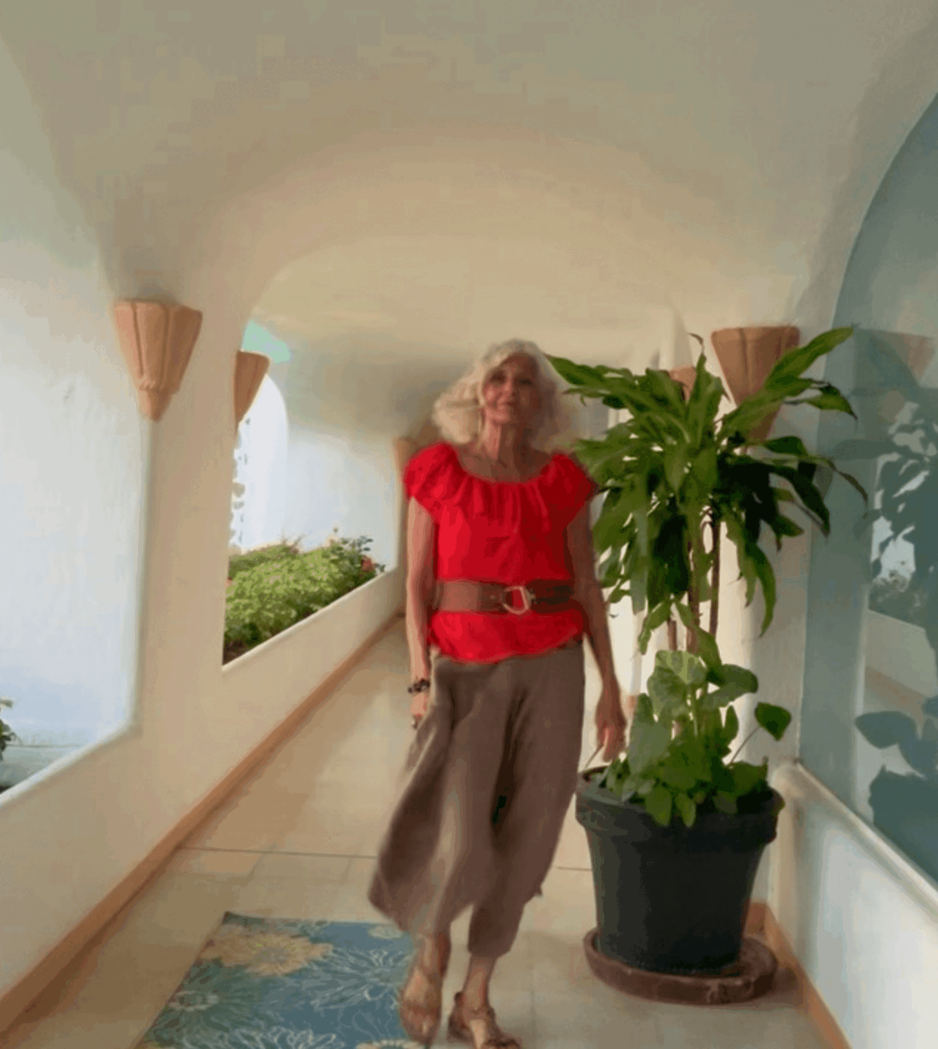 Woman in red top and beige pants walking in corridor with plant on her right side.