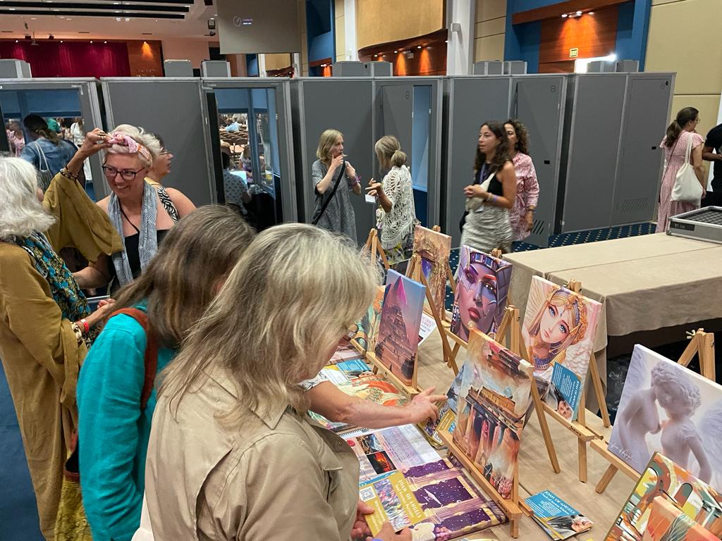 People browsing art and merchandise at an indoor market or exhibition with booths and artwork displays.