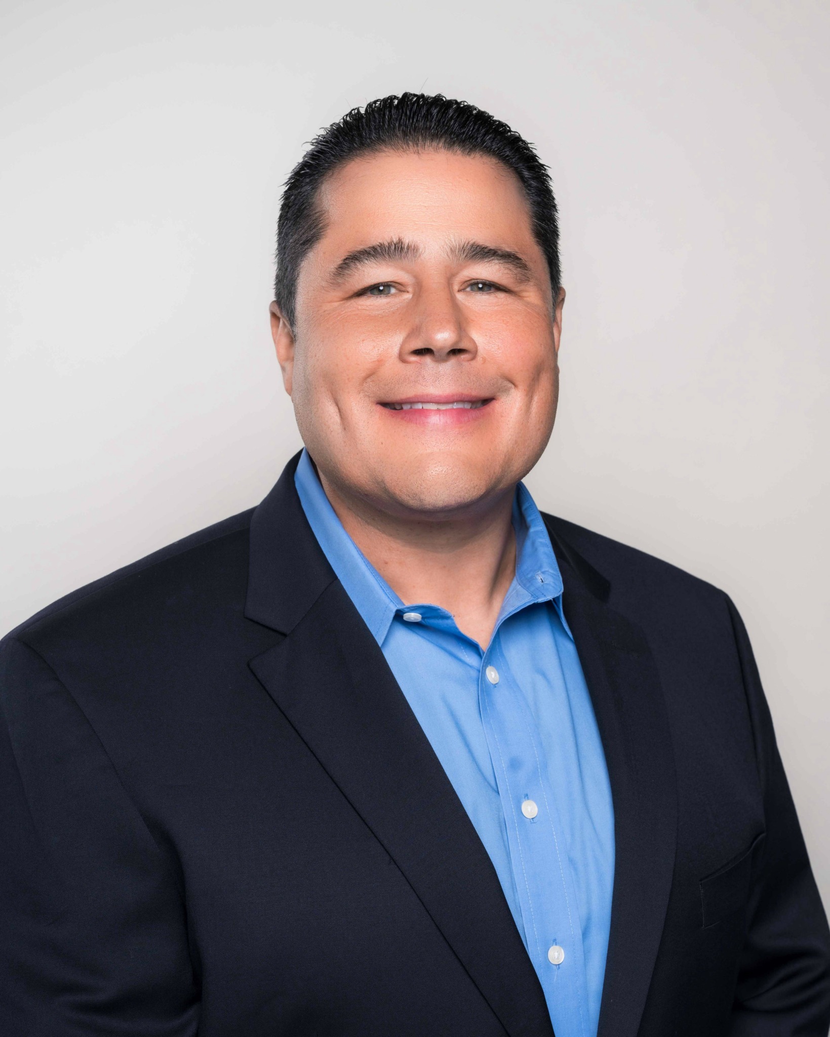 Man in a dark suit and blue shirt smiling against a plain background.