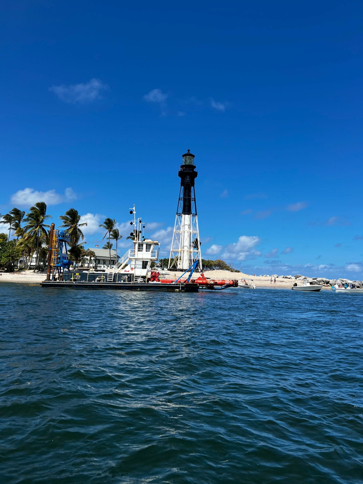 Sailboat docked at marina with blue sky and water, palm trees in background.