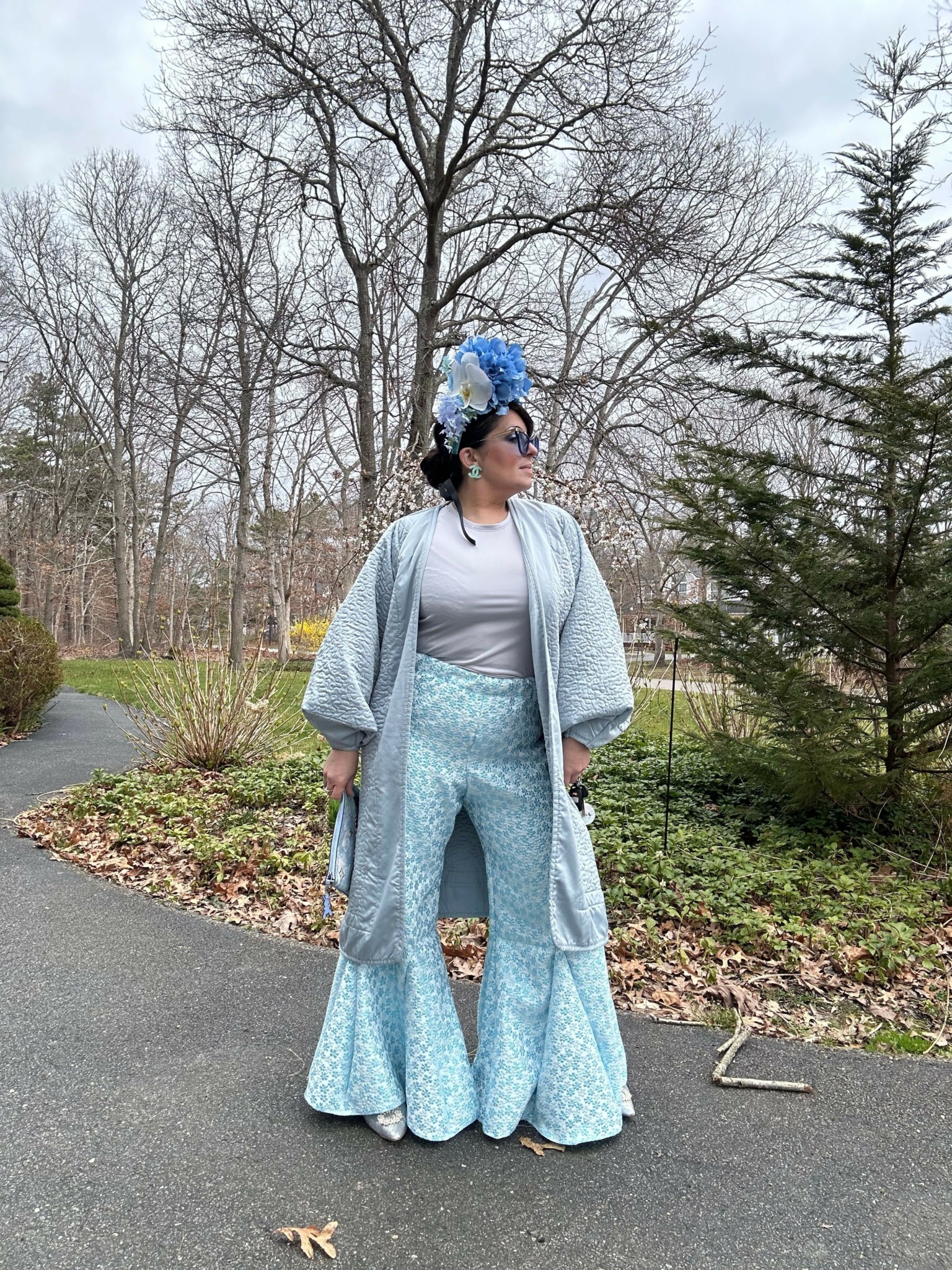 Person in light blue outfit with blue headpiece standing outdoors on paved path, trees and grass in background.