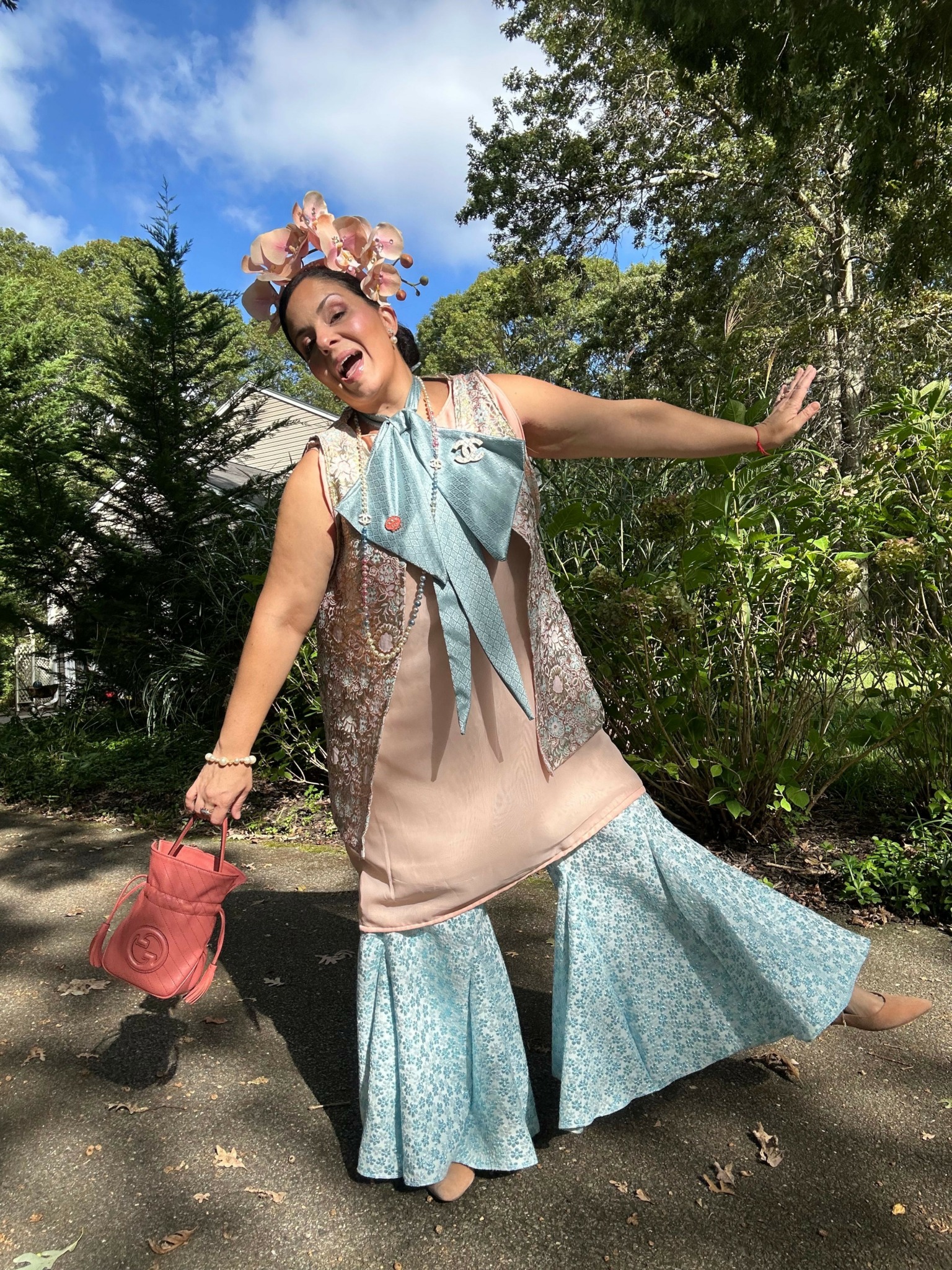 Woman in a floral dress holding a pink purse, standing outdoors with trees and blue sky in background.