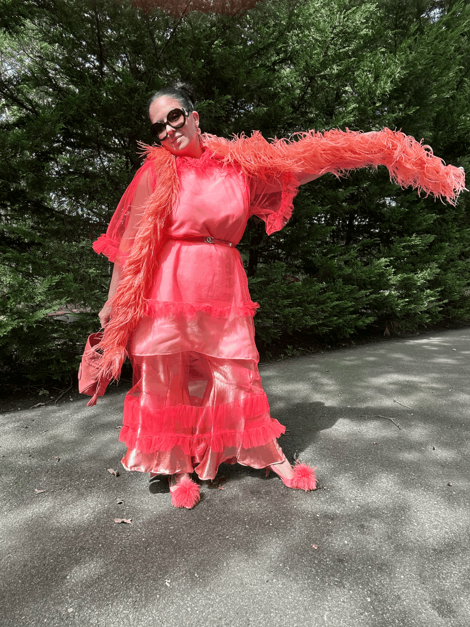 Woman in pink dress and sunglasses standing outdoors on a paved surface with green trees in background.