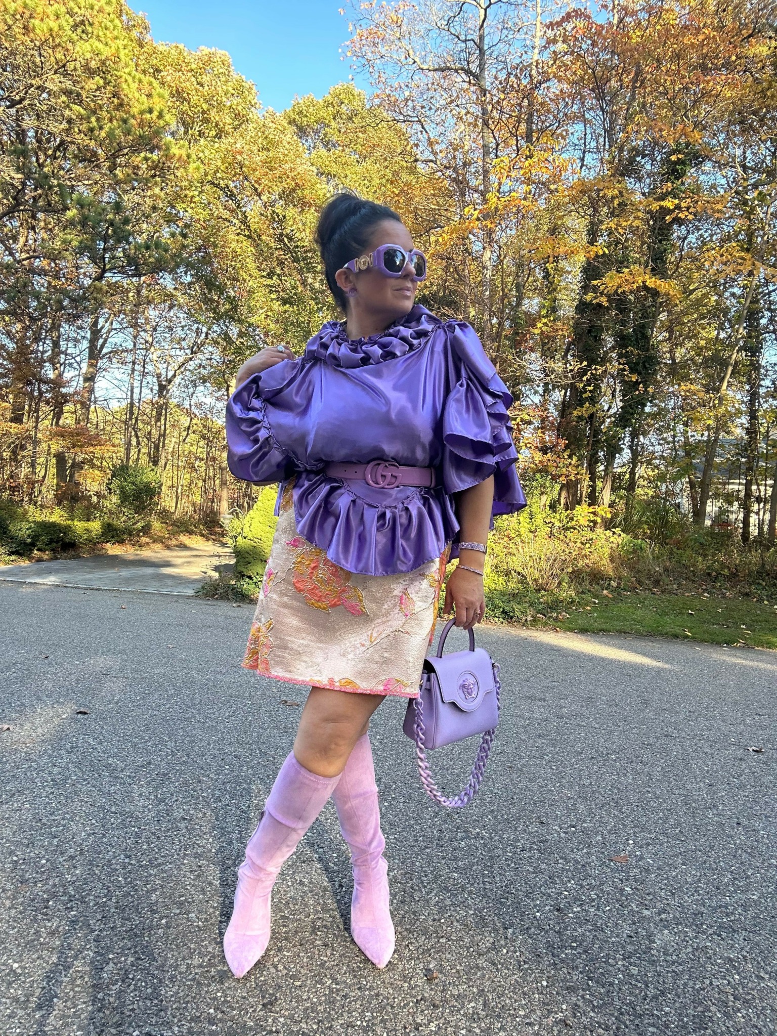 Person standing on a paved path with trees and autumn foliage in the background, wearing a purple jacket, patterned skirt, and pink boots.
