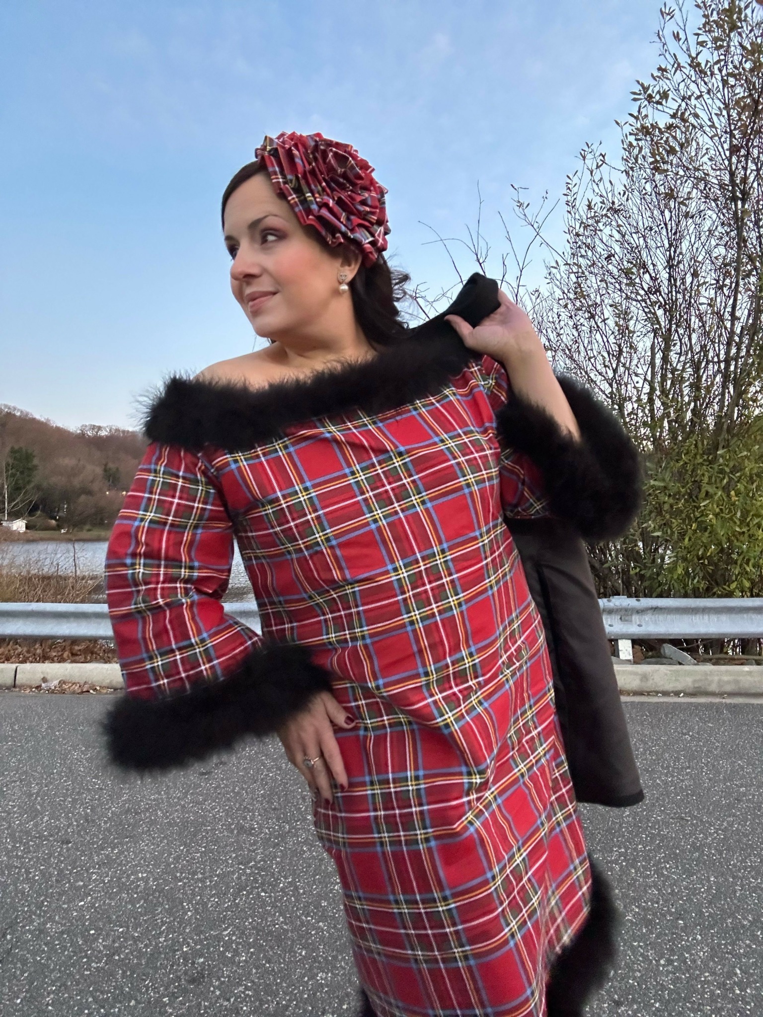 Woman in a red plaid dress with black fur trim standing outdoors on a road with trees and sky in background.
