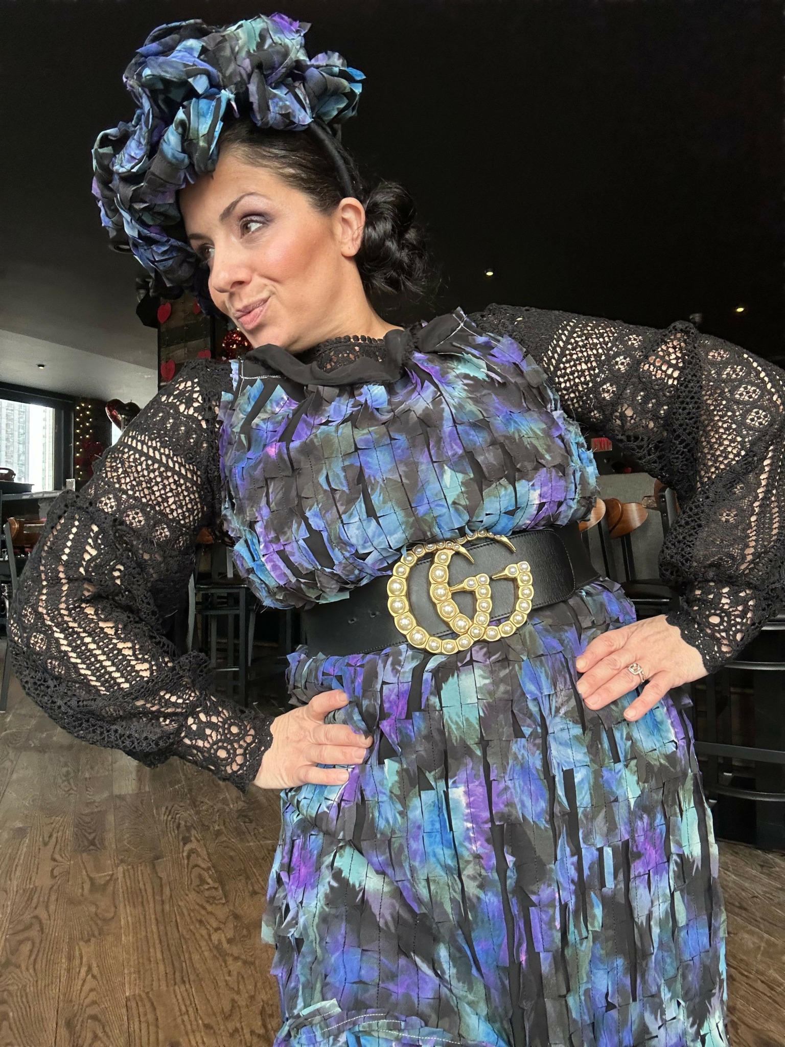 Woman in a colorful dress with lace sleeves and a matching headscarf, standing indoors on a wooden floor.