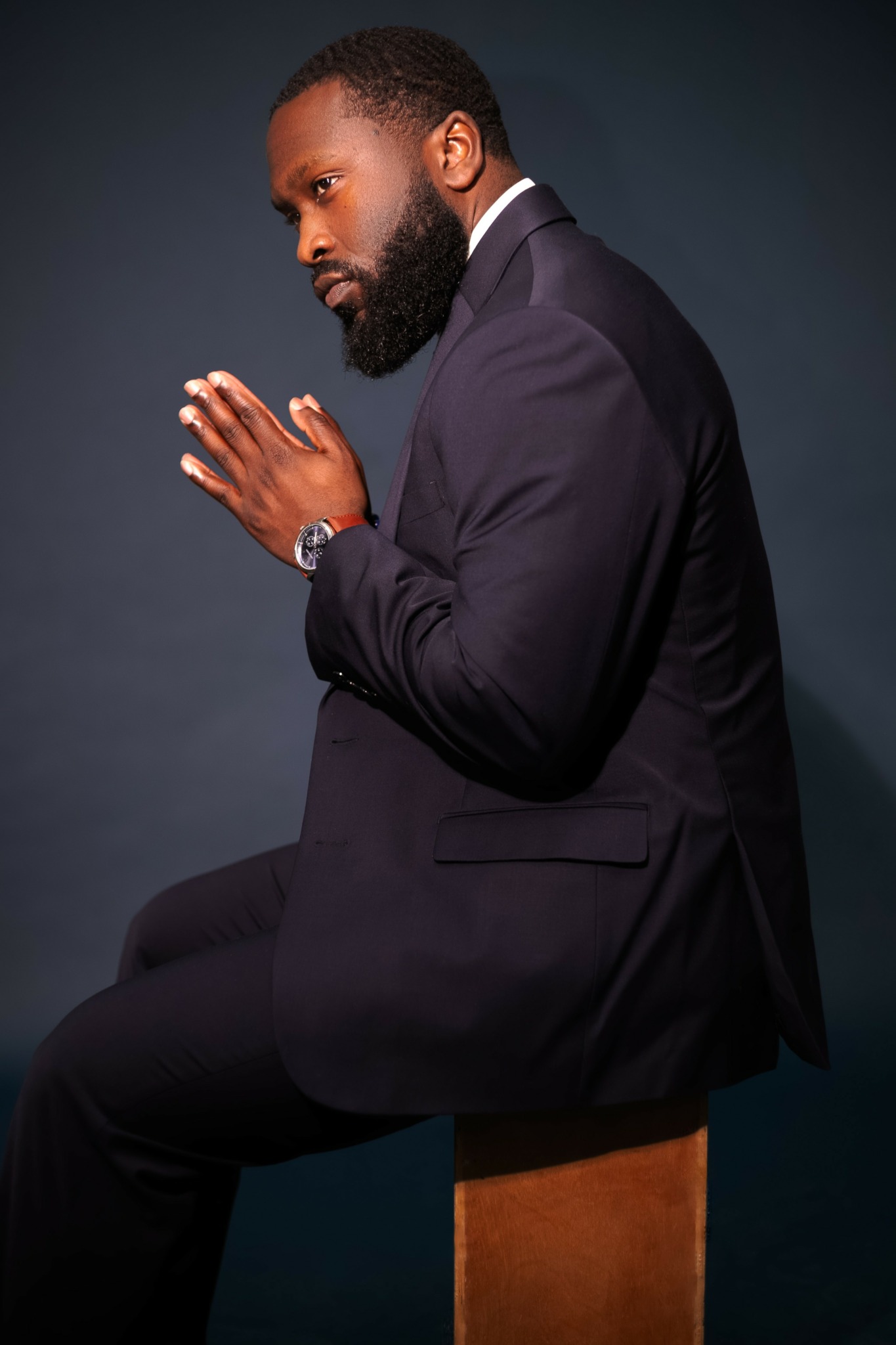 Man in suit sitting on a stool with hands pressed together, head bowed, dark background.