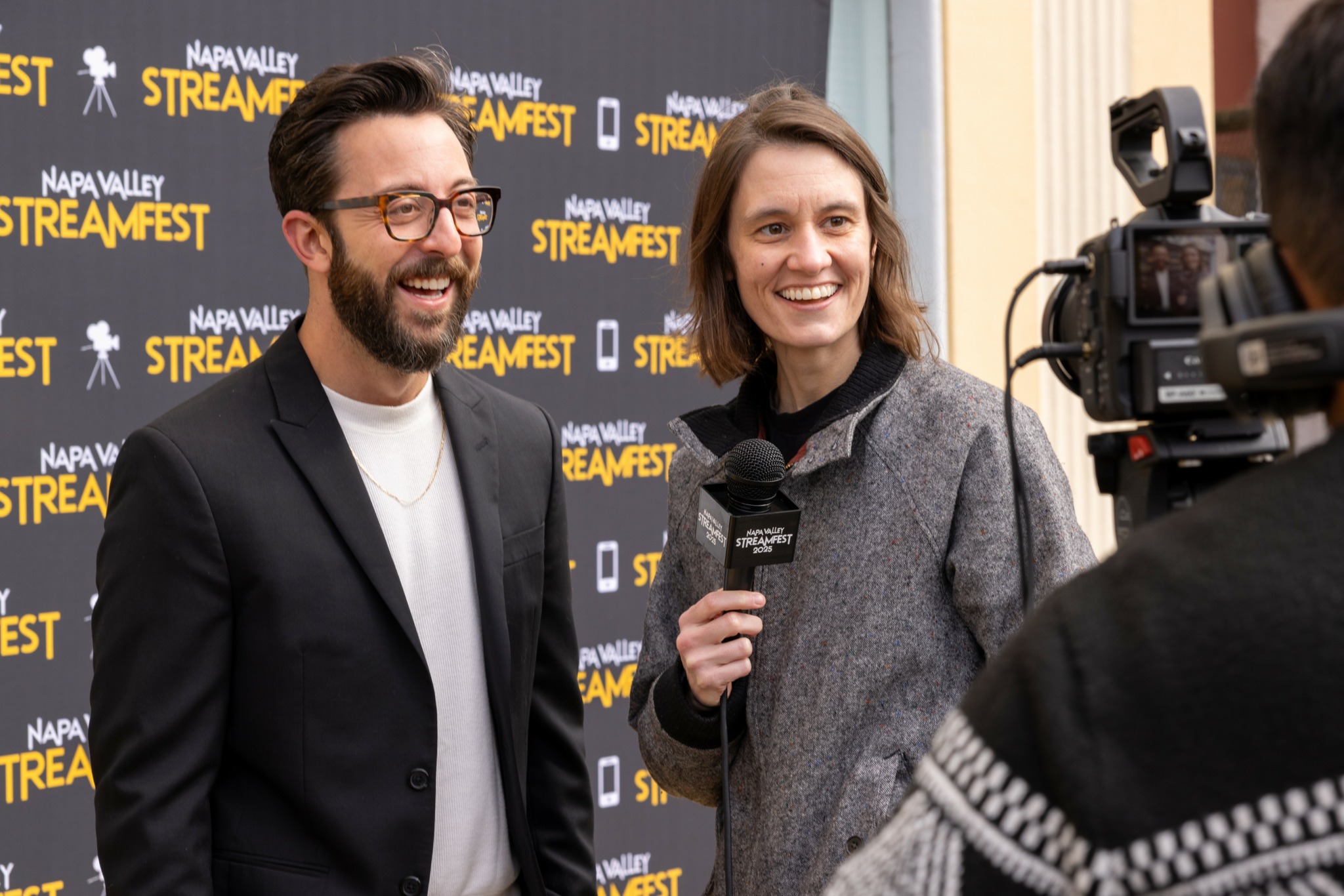 Two people being interviewed on a red carpet with a backdrop displaying event logos, smiling and talking to a camera.