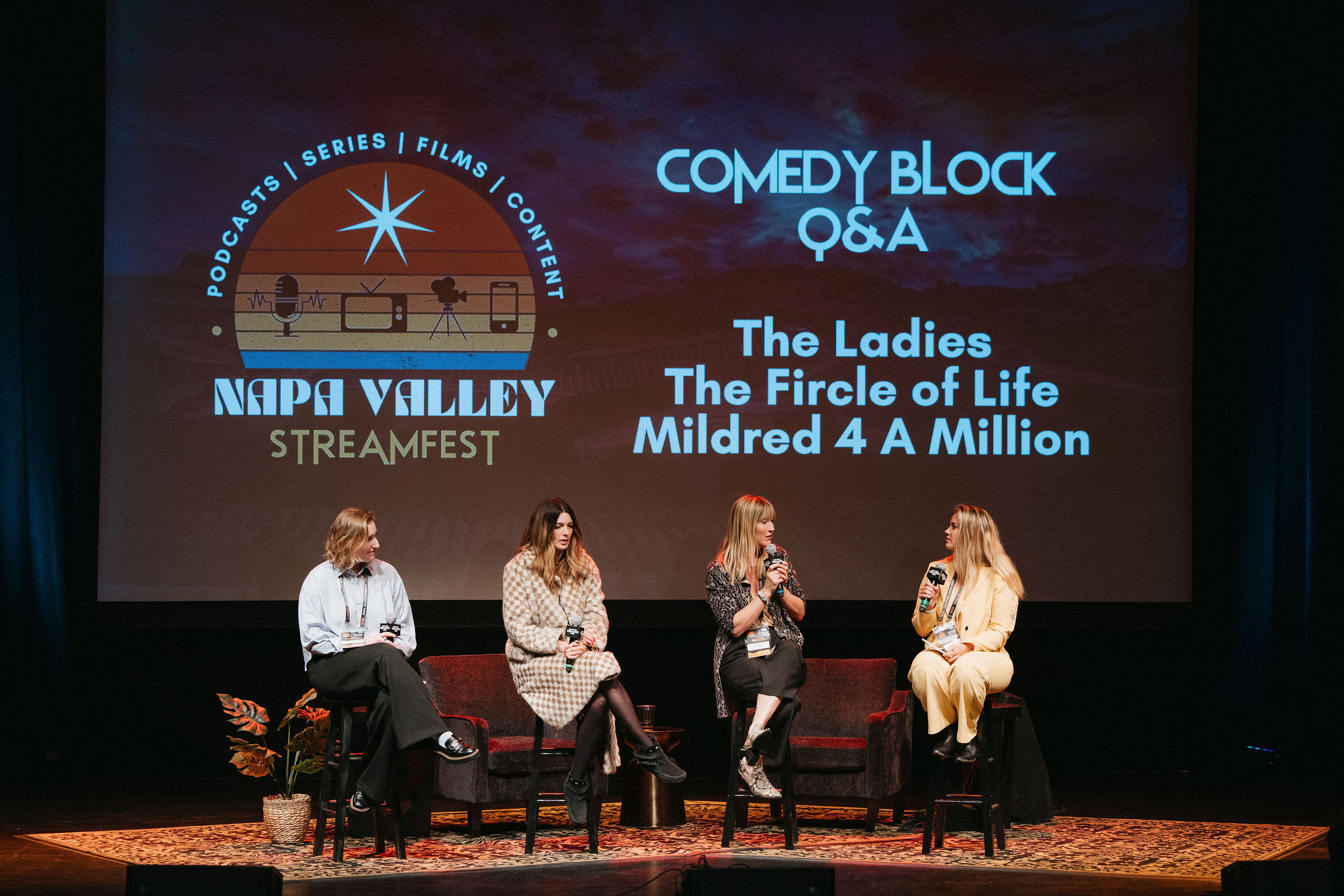 Four women seated on stage during a panel discussion with a large screen behind them displaying event information.