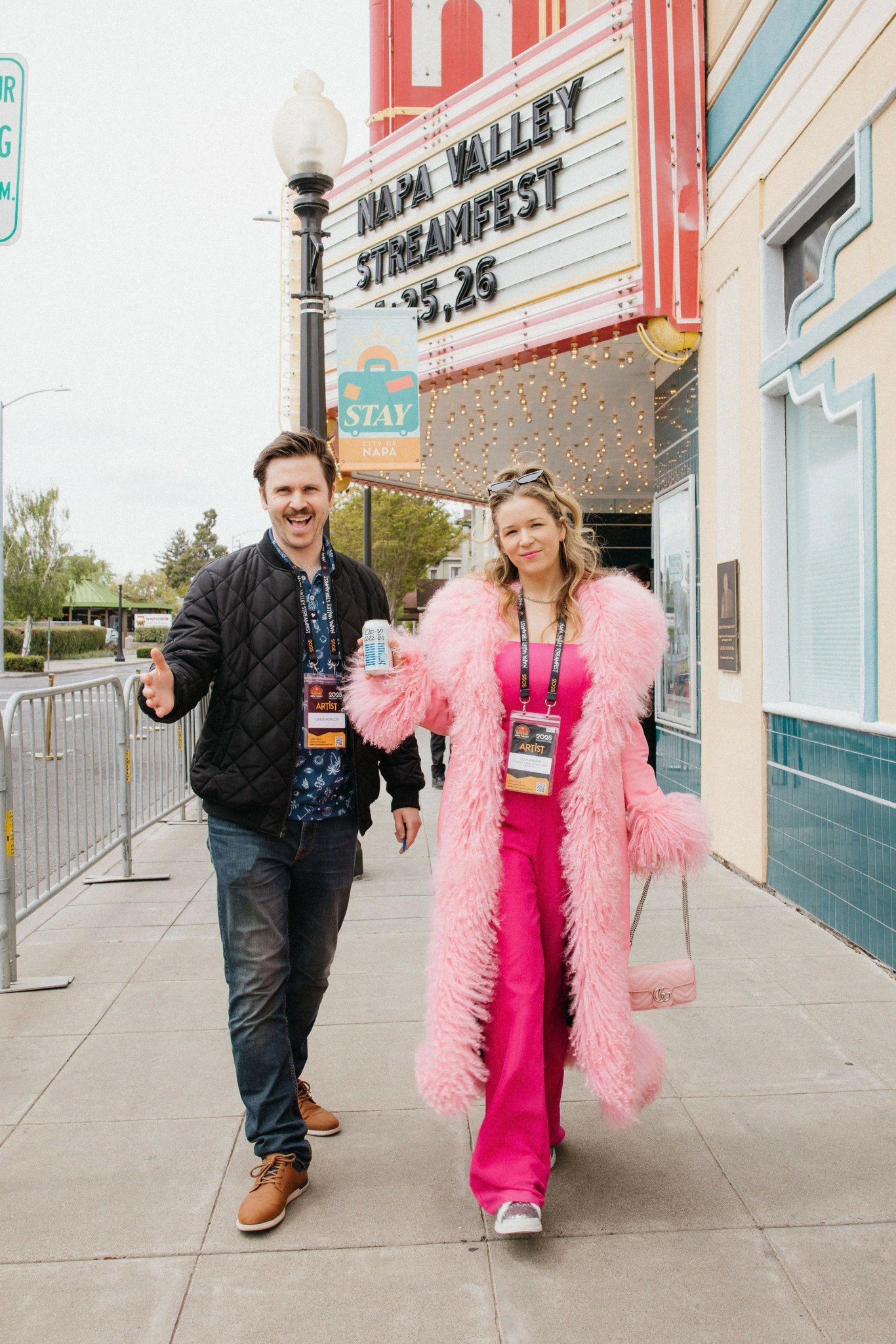 Two people walking on sidewalk in front of a theater marquee, one in pink coat, the other in dark jacket.