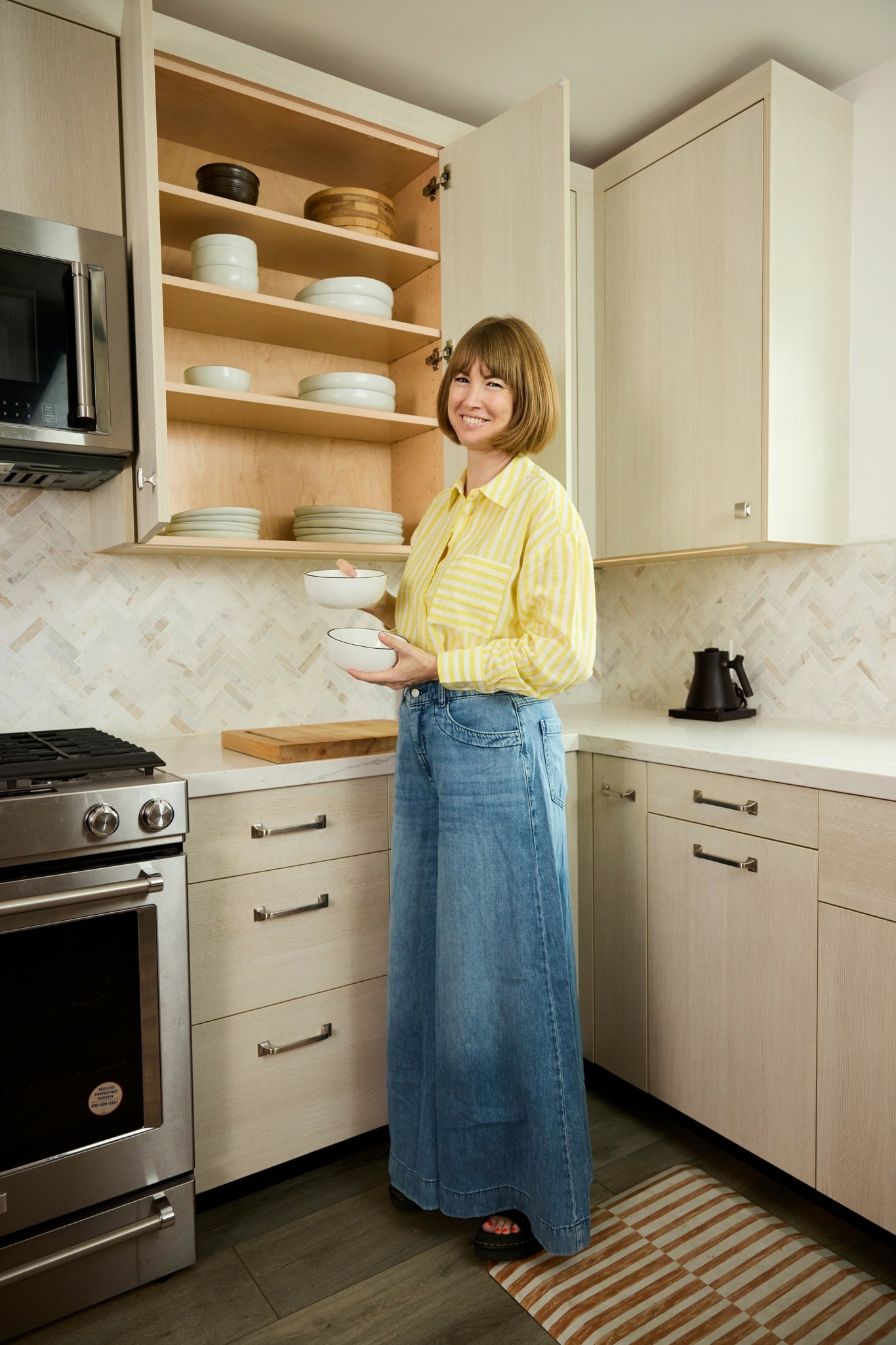 Woman in yellow shirt and long denim skirt standing in kitchen, holding a bowl, open cabinet with dishes behind her.