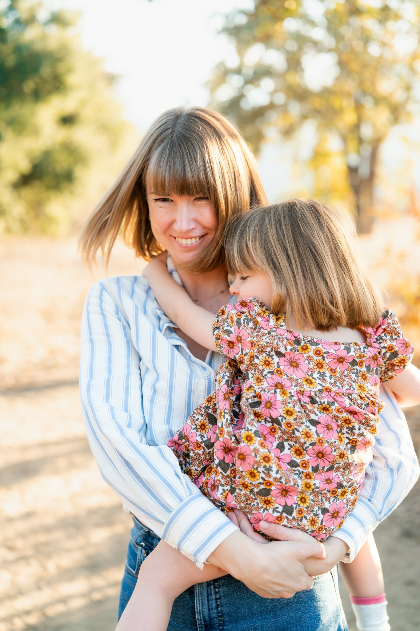 Woman holding a young girl outdoors, both smiling, with trees and sunlight in the background.