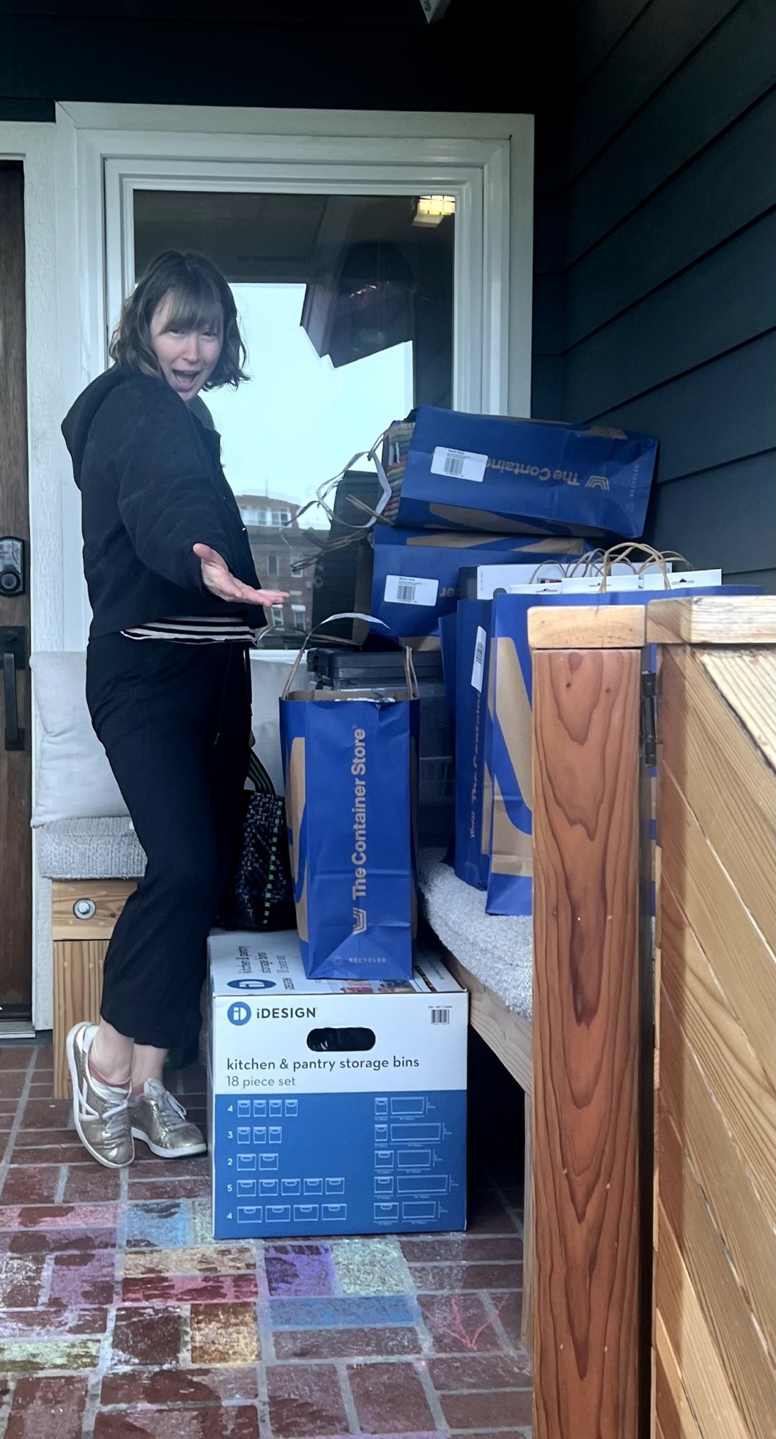 Woman standing outside near a door, holding a box, with stacked boxes around her and a wooden fence nearby.