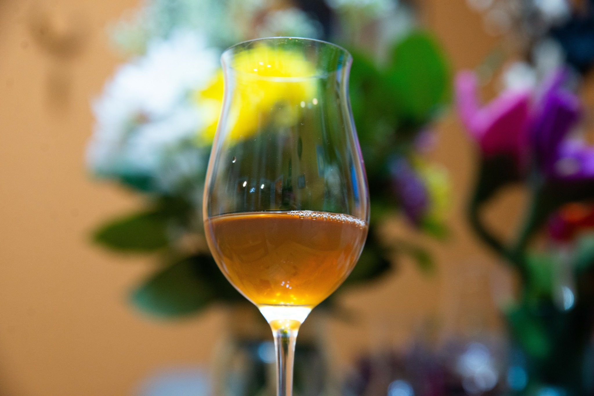 A tall glass with a yellow flower inside, filled with a pinkish beverage, in front of a blurred floral background.