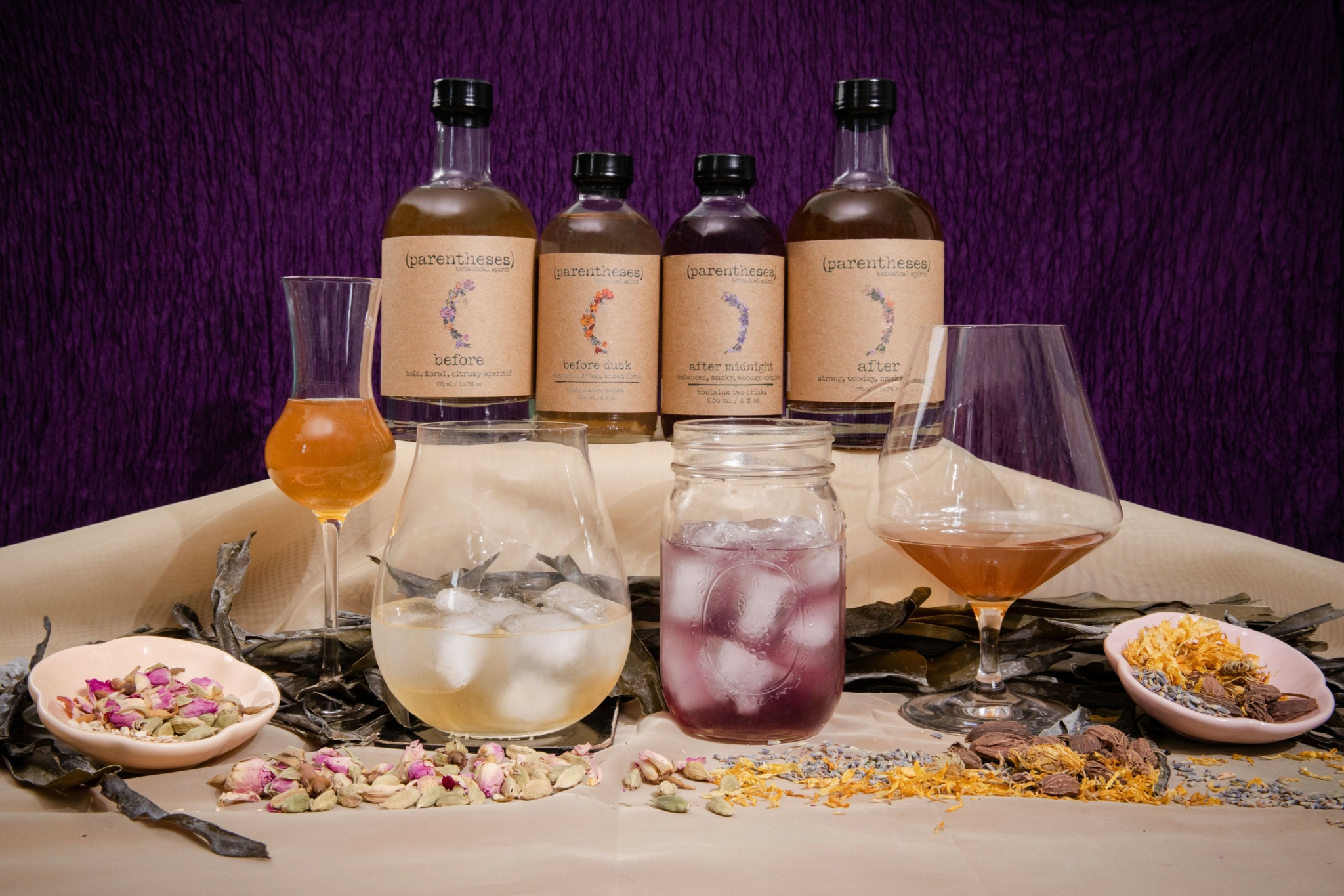Four bottles of whiskey, glasses with drinks, and bowls with snacks on a table against a purple background.