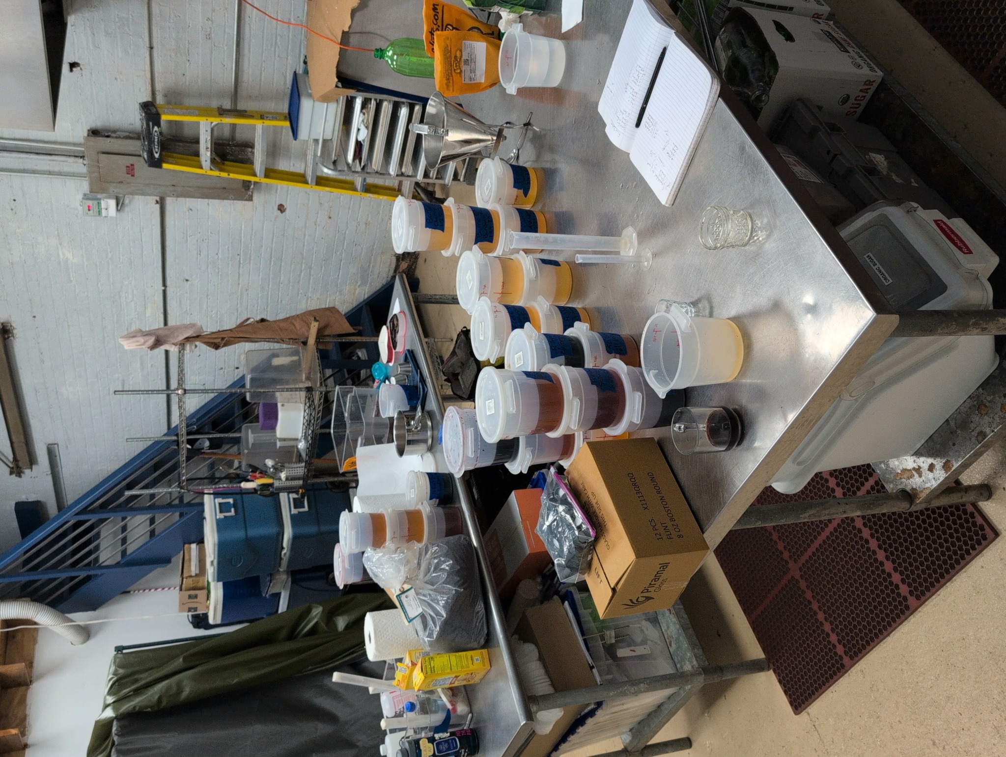 Table with numerous cups, bottles, and boxes in a cluttered industrial setting, with a person partially visible in the background.