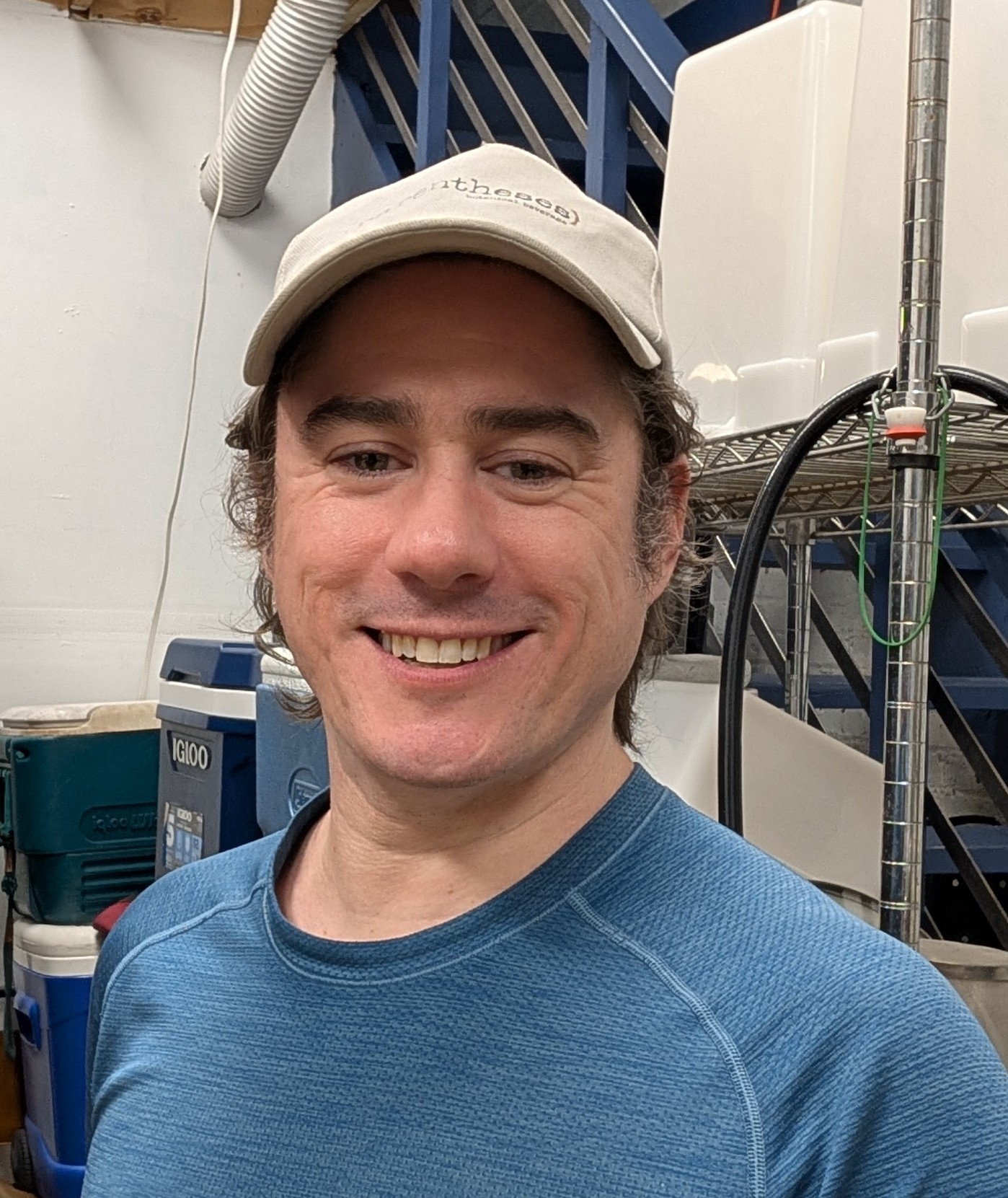 Smiling man wearing a beige cap and blue shirt indoors with equipment and stairs in background.