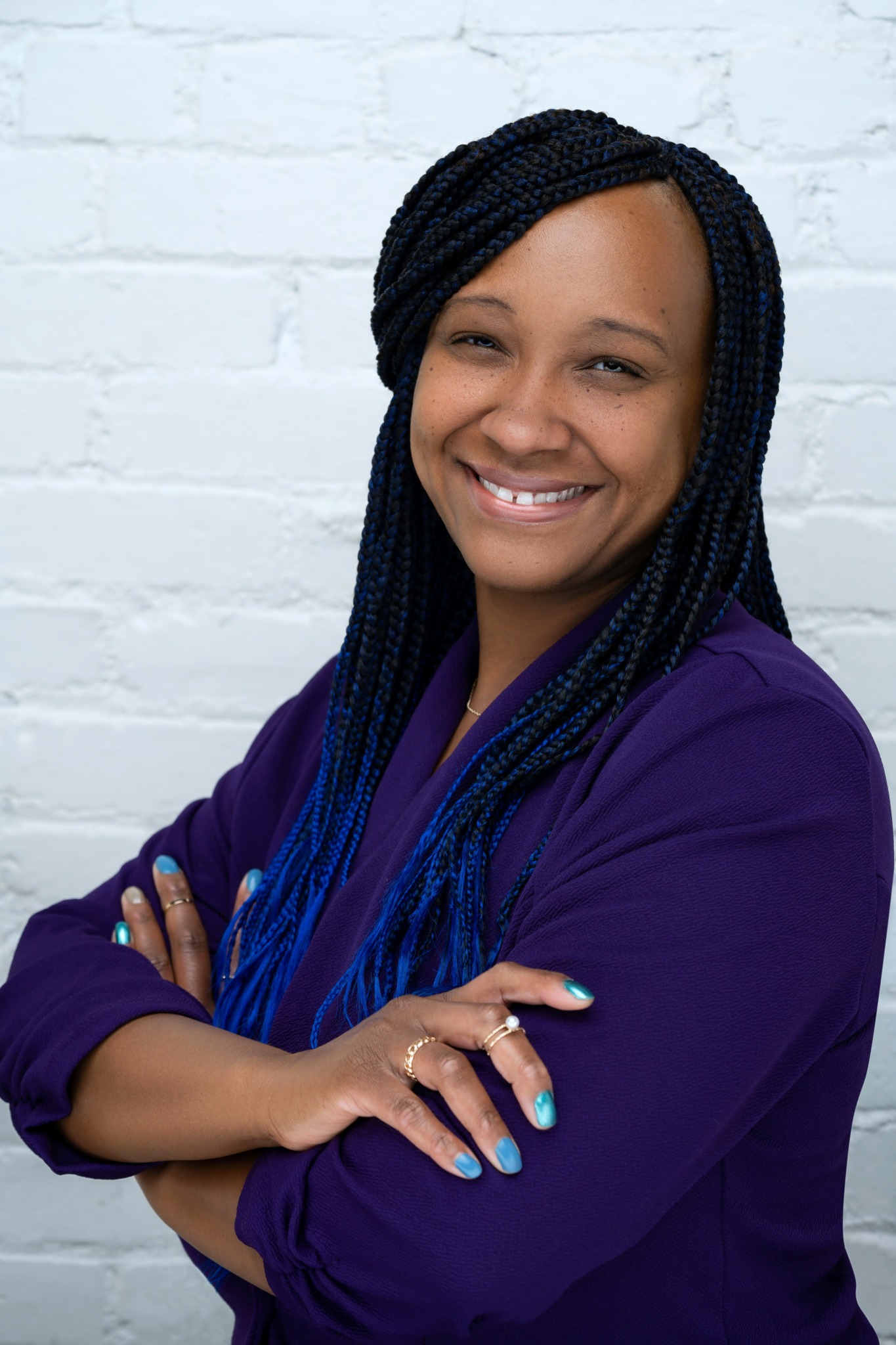 Smiling woman with braided hair and purple top standing against white brick wall.