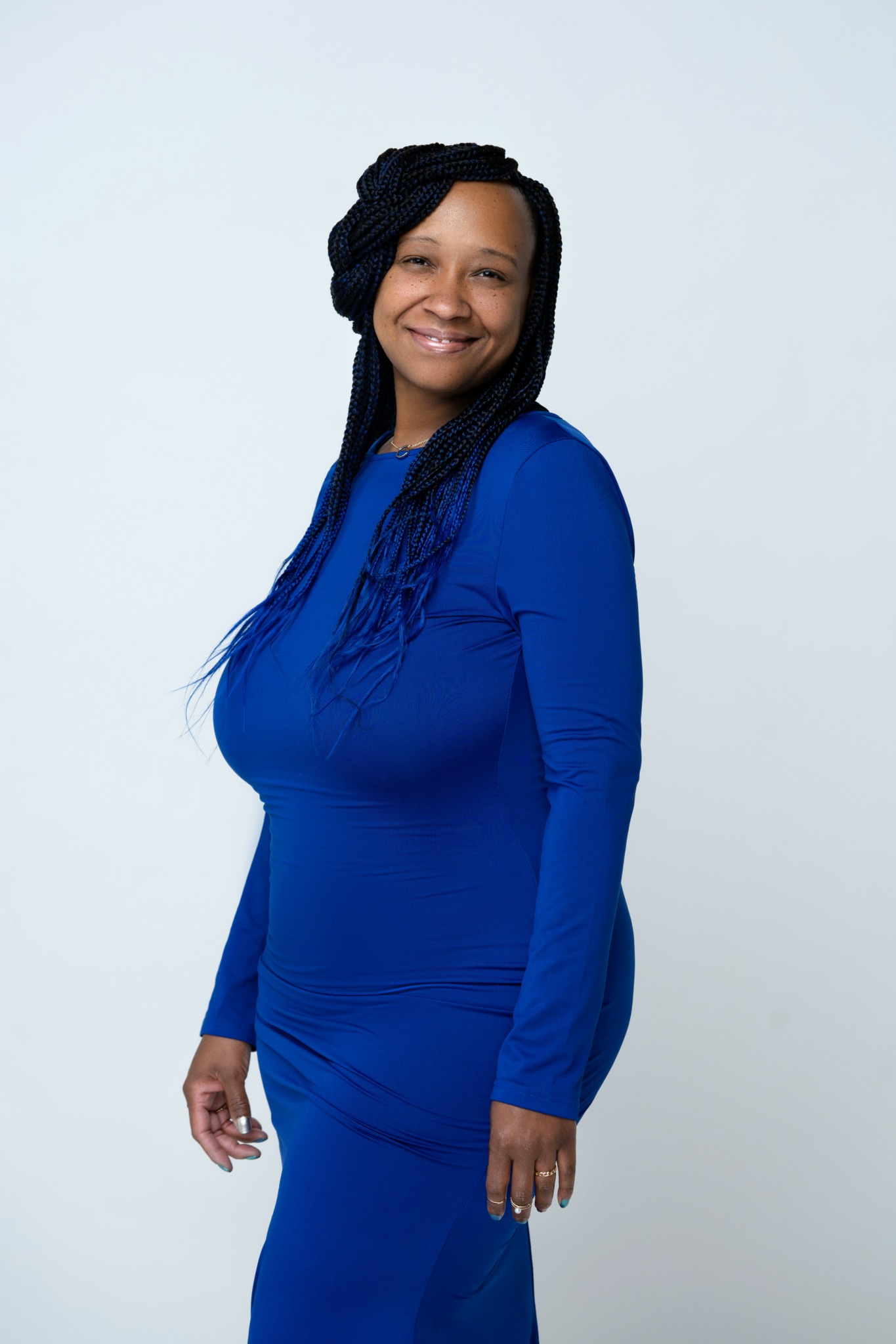 Woman with long braided hair wearing a blue dress, smiling, standing against a plain background.