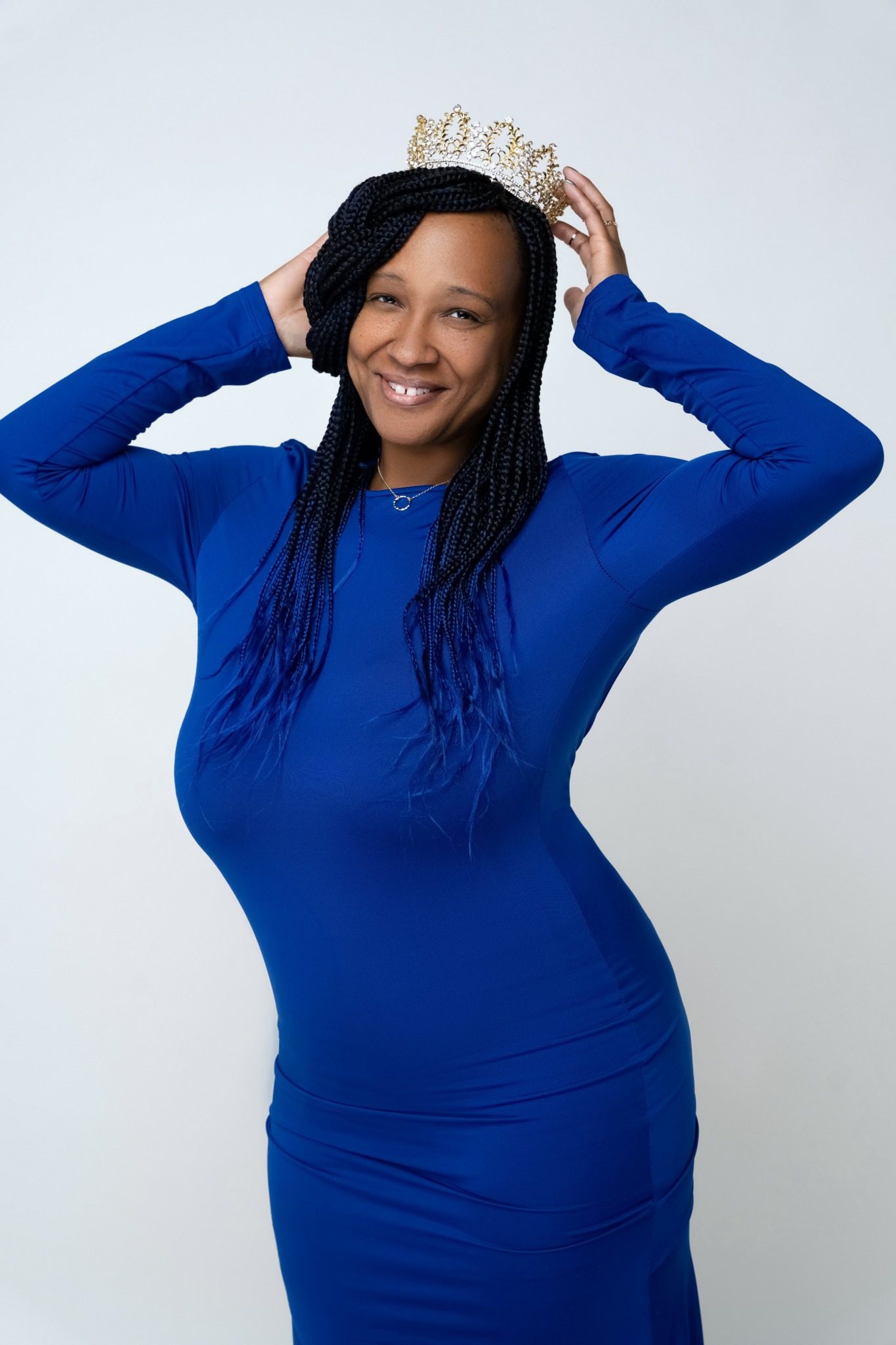 Woman wearing a blue dress and crown, smiling, holding crown with both hands, plain light background.