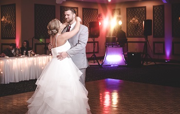 Couple dancing in a decorated reception hall with a DJ booth and colorful lighting.