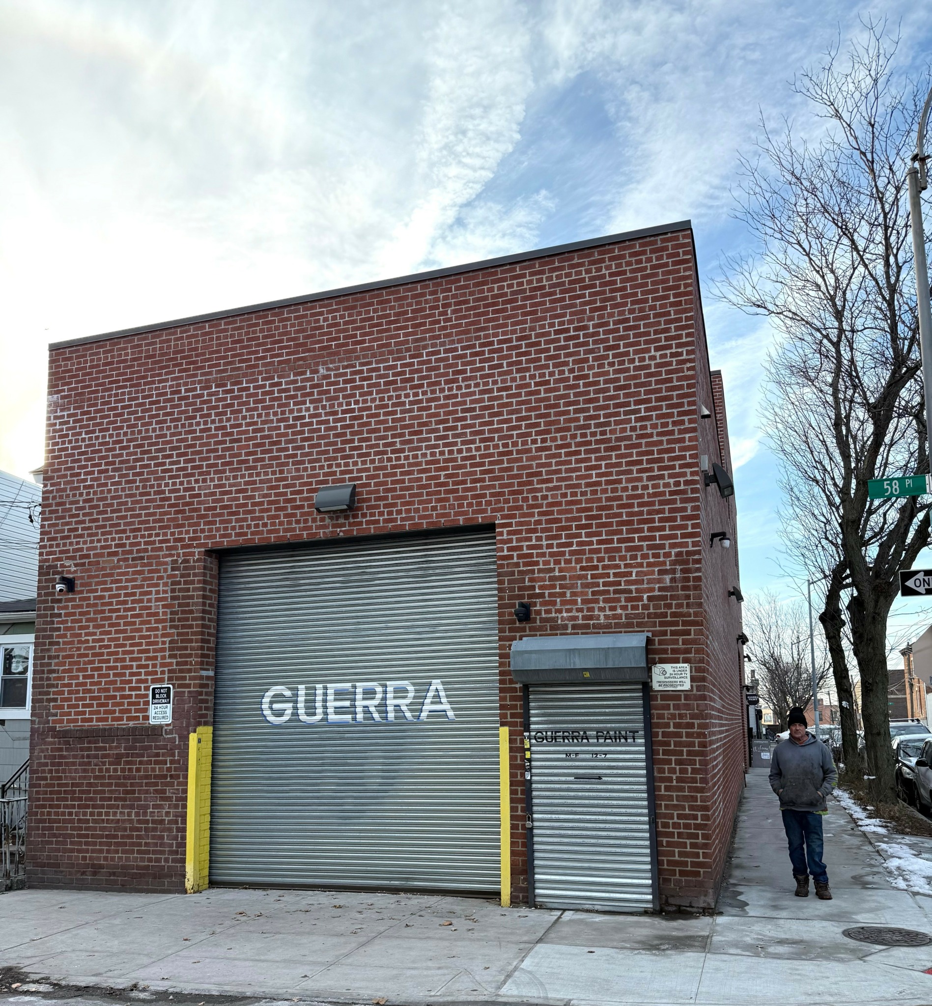 Brick building with a large metal garage door and a smaller door, with a person walking nearby and leafless trees.