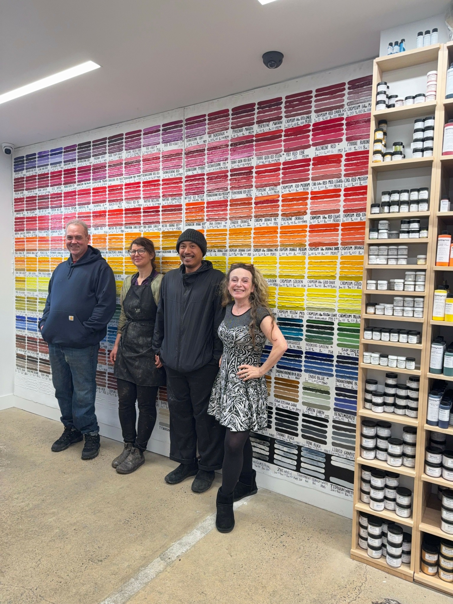 Four people standing in front of a wall of colorful thread spools in a store, with shelves above and below.