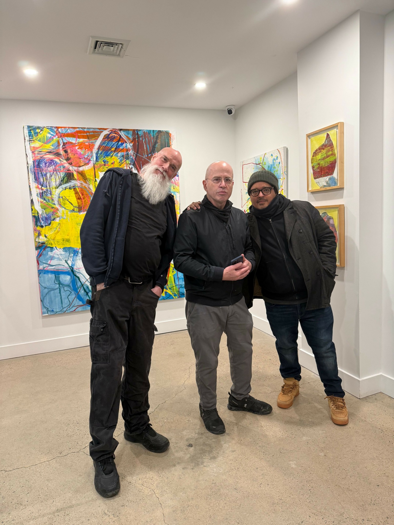 Three people standing in an art gallery with colorful paintings on the wall behind them.
