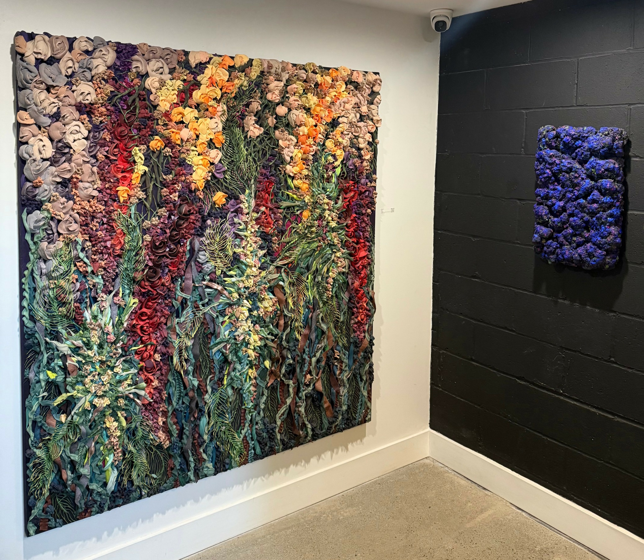 Two textured floral art pieces on white and black walls in a gallery space.