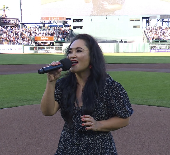 Woman singing into microphone on sports field with crowd in background.