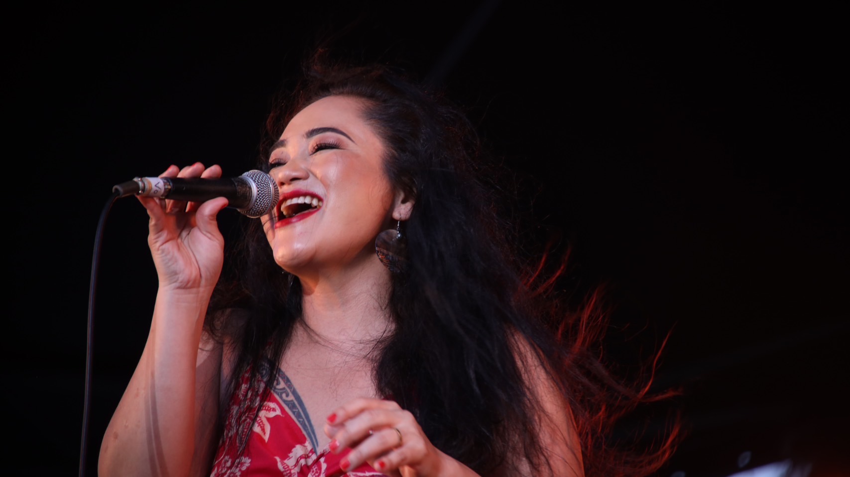 Woman singing into a microphone, smiling, with long dark hair and a red patterned top.