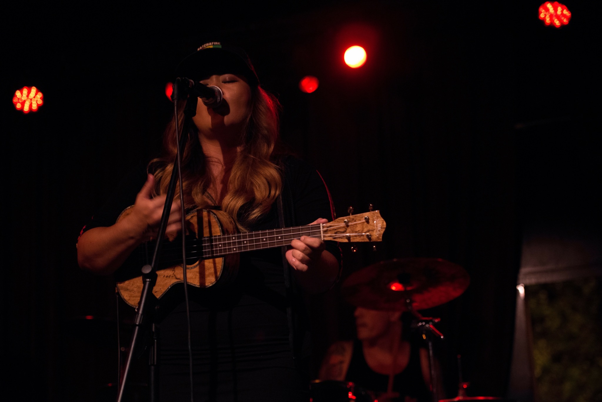 Female musician singing into a microphone while playing a ukulele on stage with red lighting.