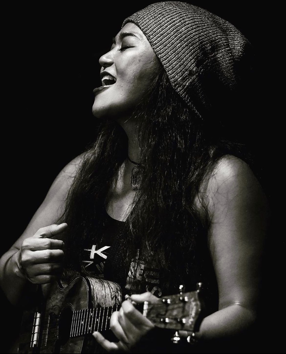 Woman wearing a beanie, smiling, playing guitar, in black and white, side profile, dark background.