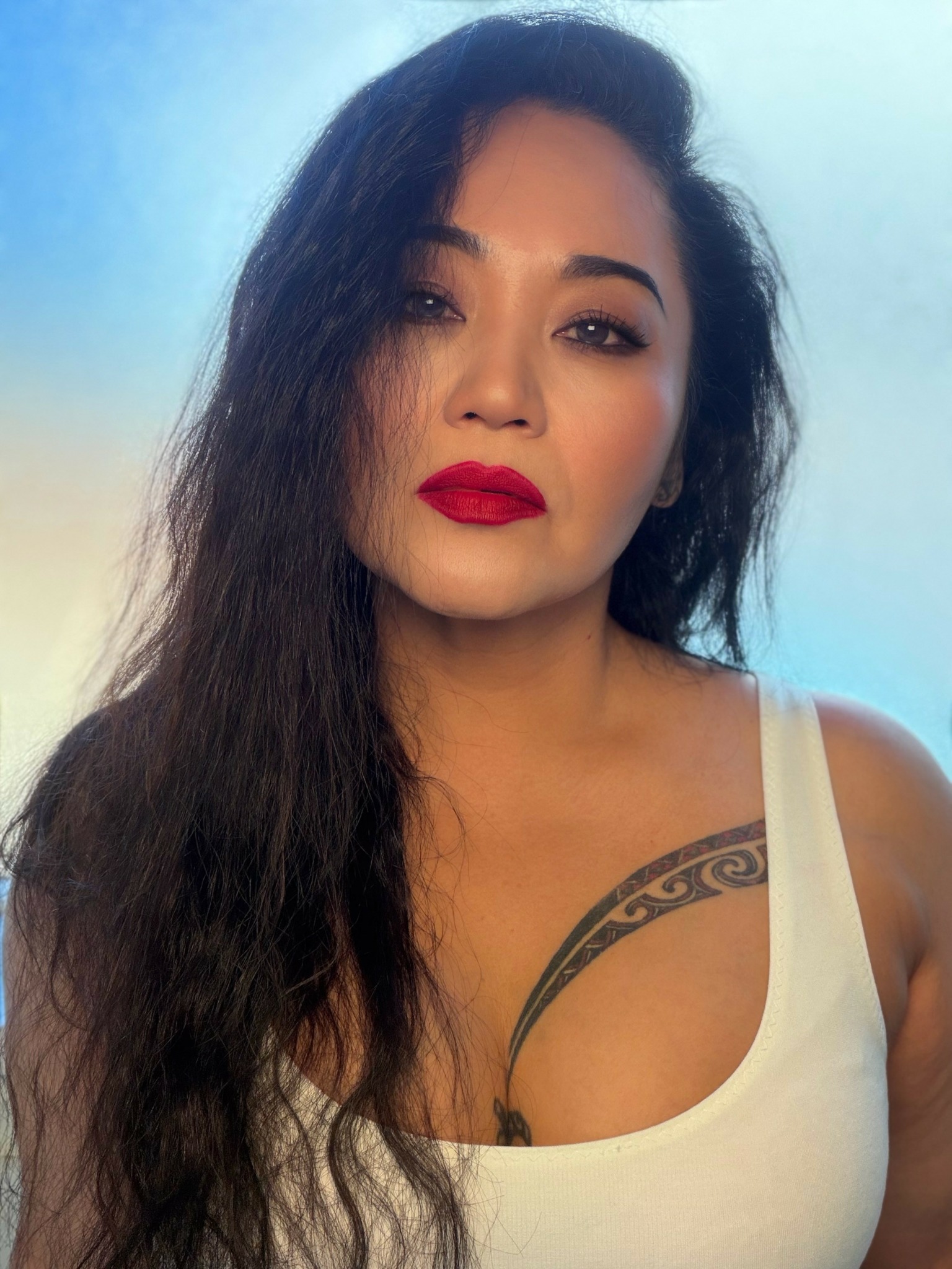 Woman with long dark hair, red lipstick, wearing a white top, tattoo visible on shoulder.