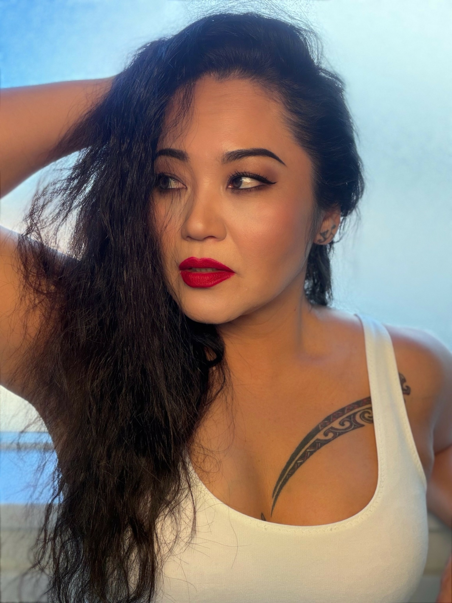 Woman with long dark hair, red lipstick, wearing a white tank top, looking to the side.