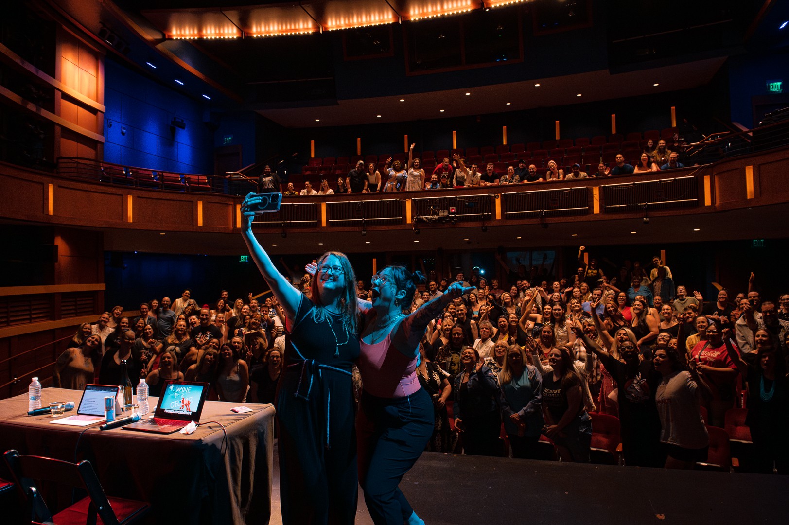 Performer on stage holding a microphone, audience clapping and watching in a large theater with multiple levels.