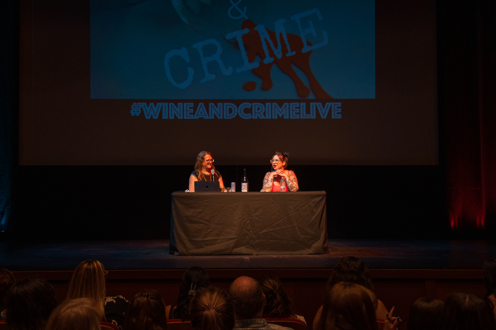 Two women sit at a table on stage, speaking to an audience in a theater setting.