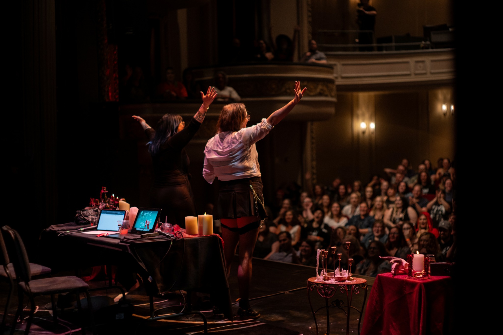 Two performers on stage with arms raised, facing an audience in a theater with balcony, tables, and candles.