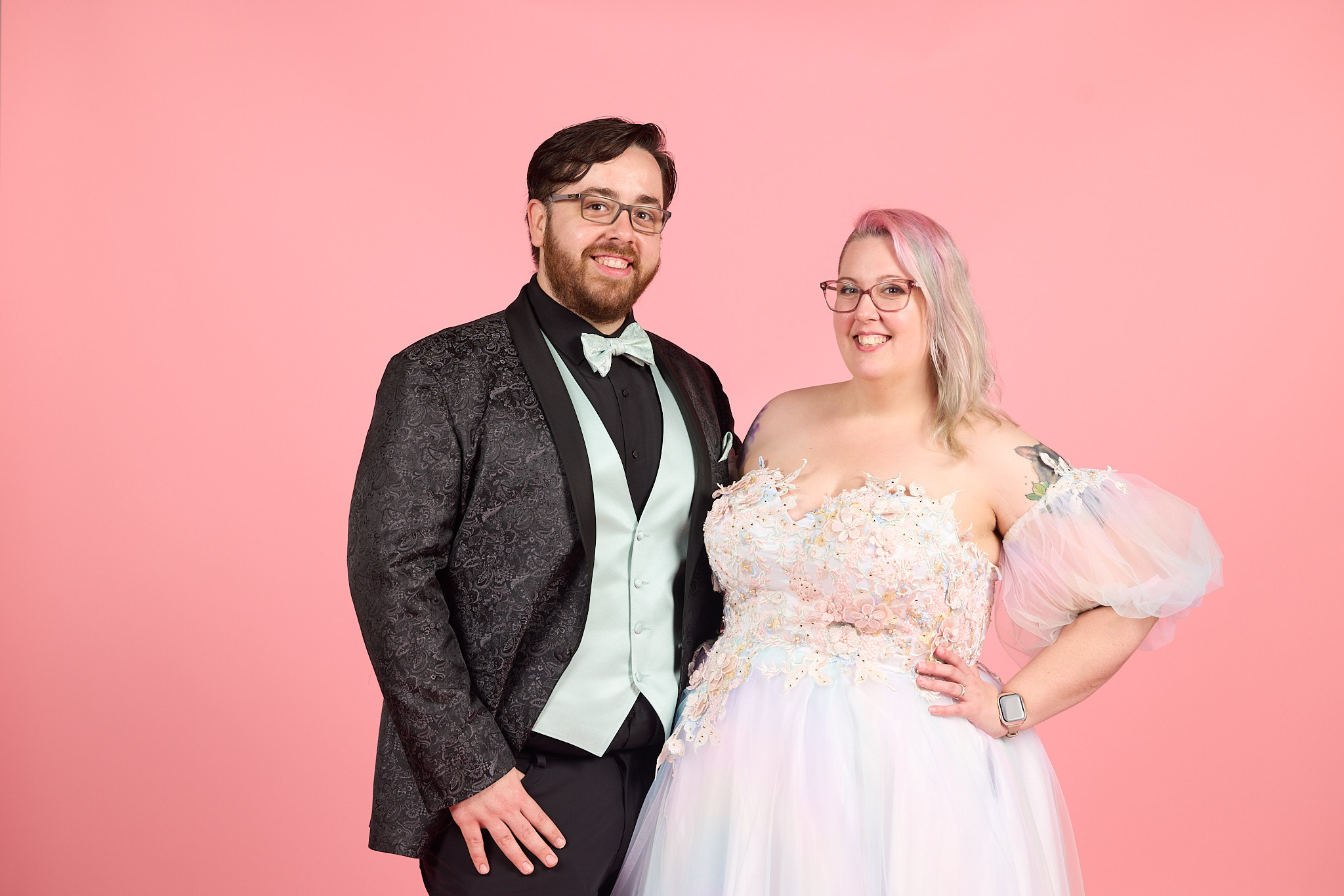 A smiling couple in formal attire stands against a pink background, the man in a tuxedo and the woman in a wedding dress.