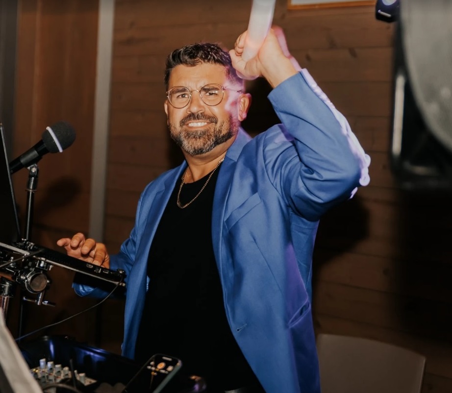 Man with glasses and beard smiling, holding a white object, standing behind DJ equipment in a wooden room.