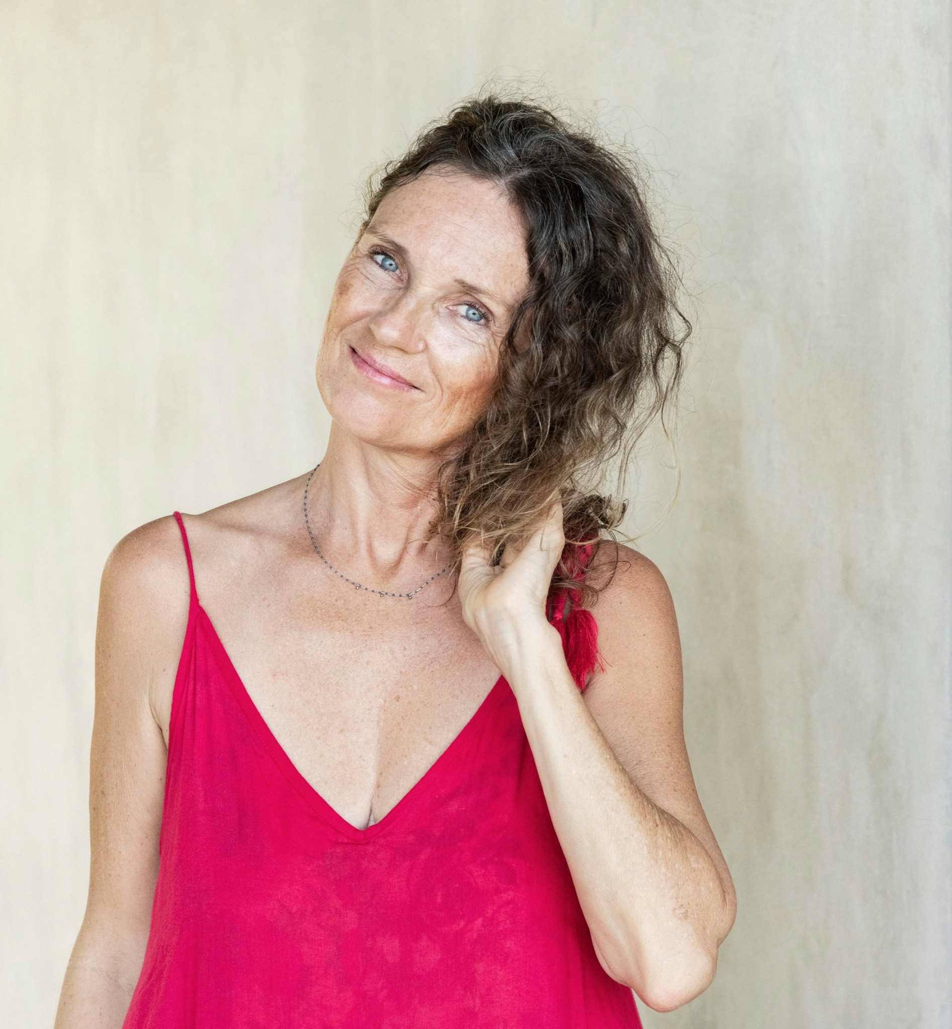 Woman with curly hair wearing a red spaghetti strap top, smiling gently, touching her hair.