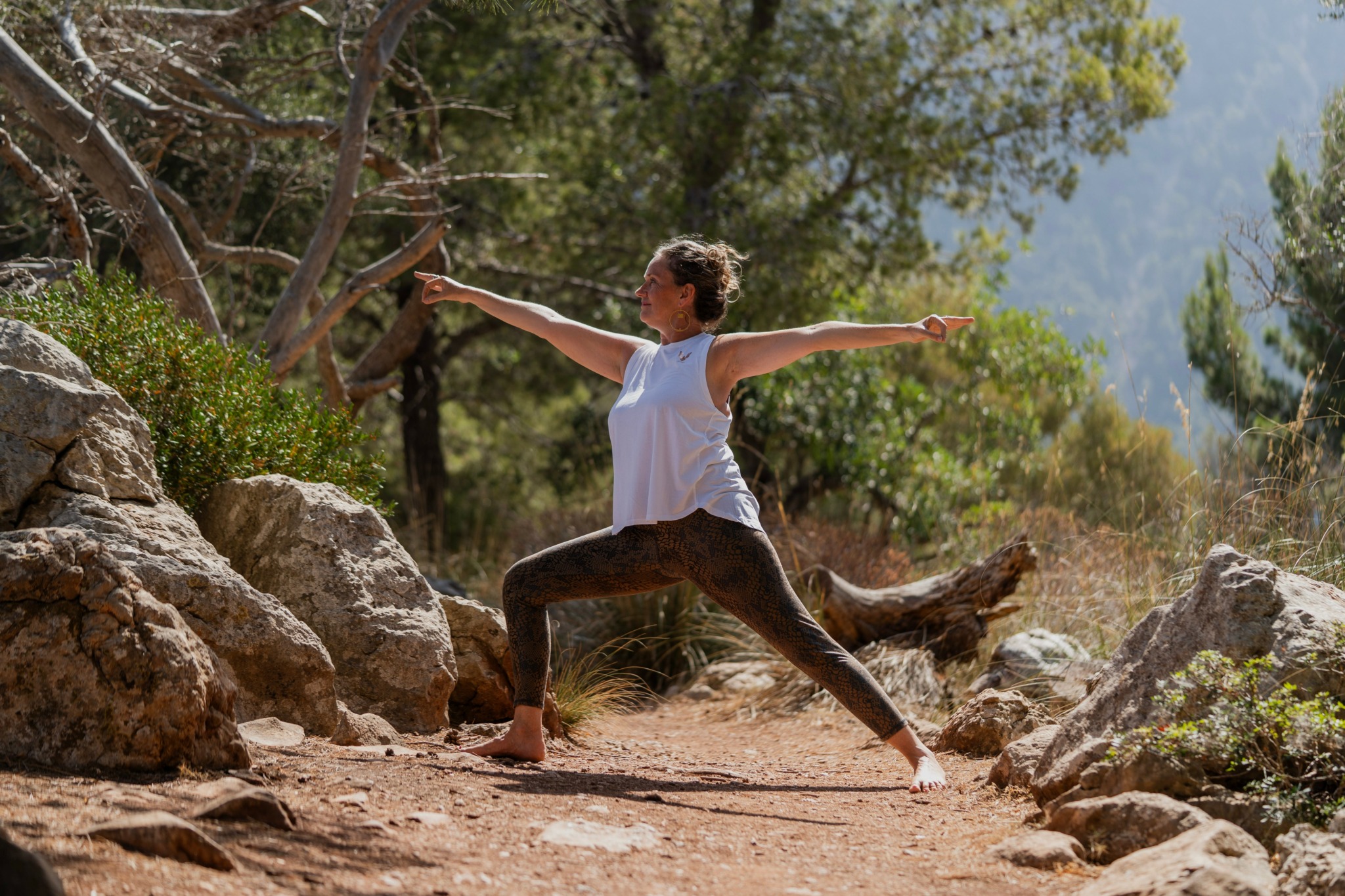 Person practicing yoga outdoors on rocky terrain with trees and mountains in background.