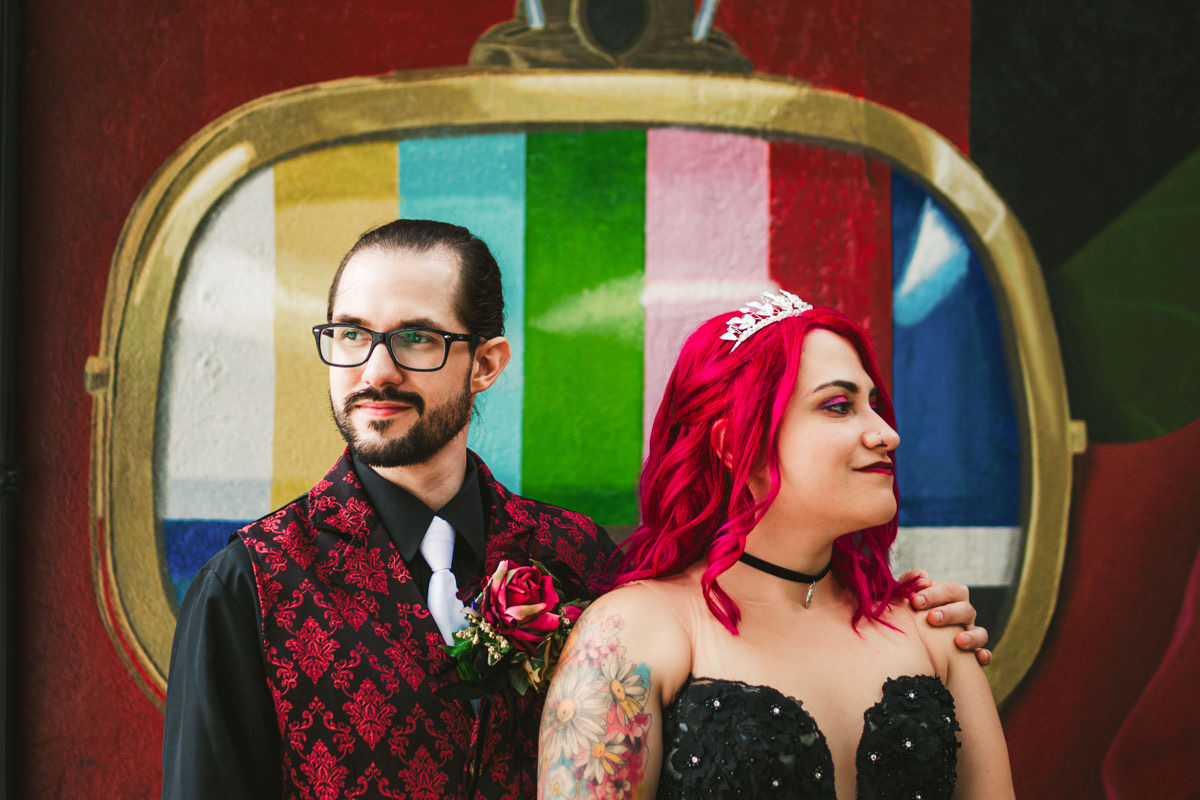 A man with glasses and a woman with red hair pose together in front of a colorful television screen background.