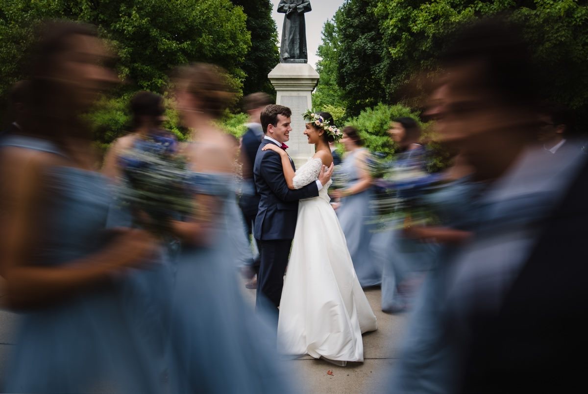 Bride and groom dance in center, surrounded by blurred wedding guests outdoors with trees and statue background.